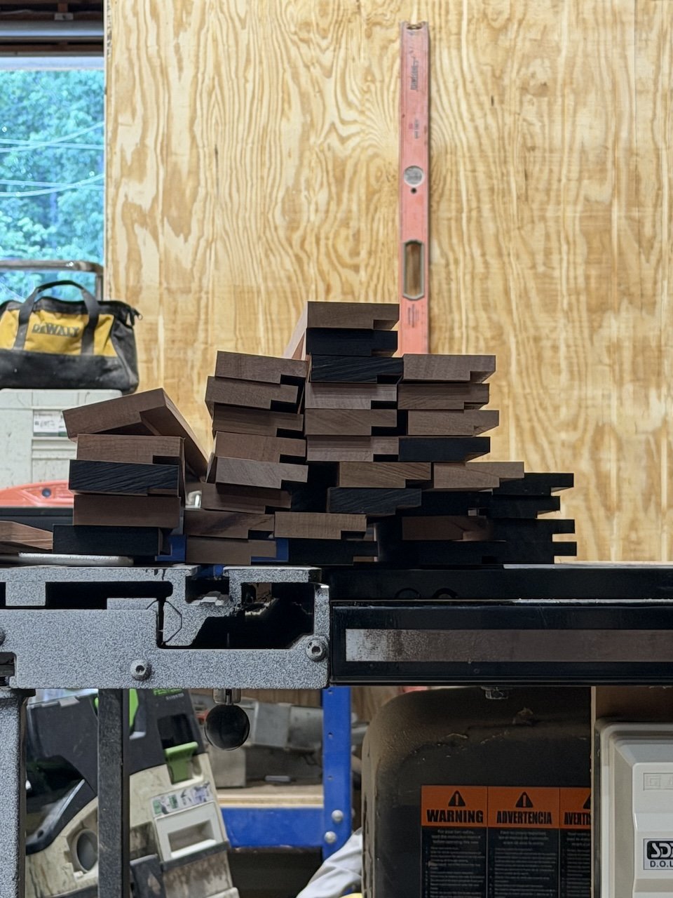 Stacked pieces of wooden molding or trim on a workshop table in front of a wooden wall, with a level tool standing vertically on the wall.