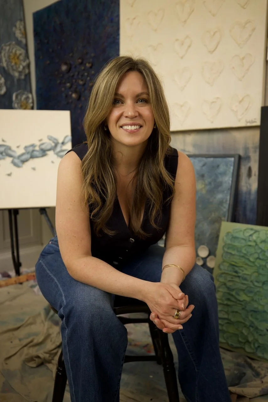 A smiling woman with wavy brown hair, wearing a black sleeveless top and blue jeans, seated in an art studio with abstract paintings and canvases in the background.