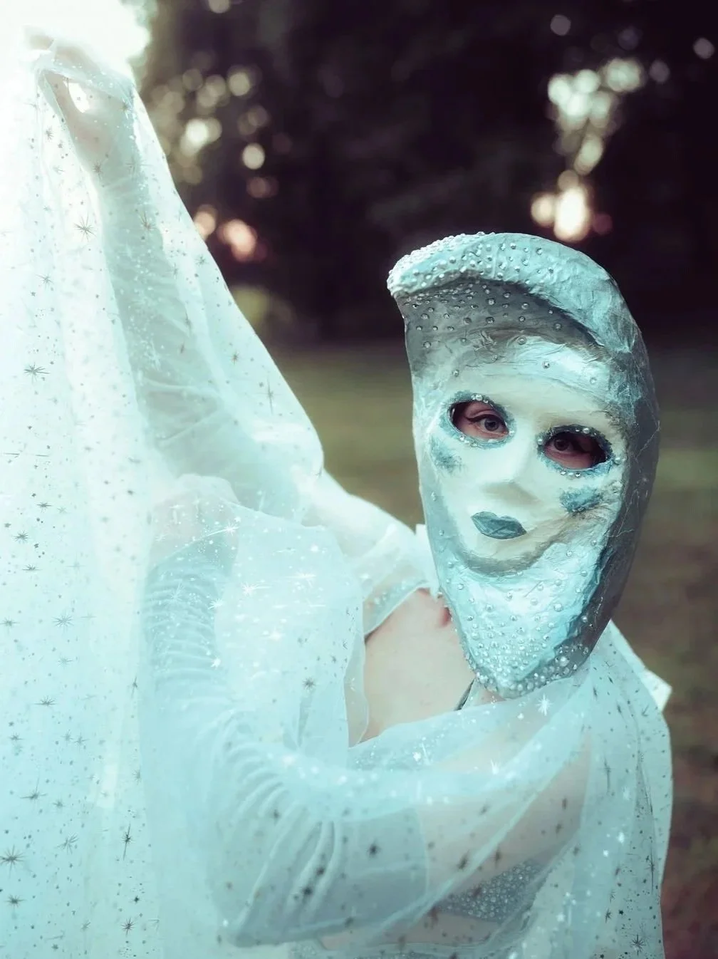 Person wearing a silver mask with eye and lip openings, decorated with small bumps, and a flowing white fabric with star patterns, outdoors during sunset.