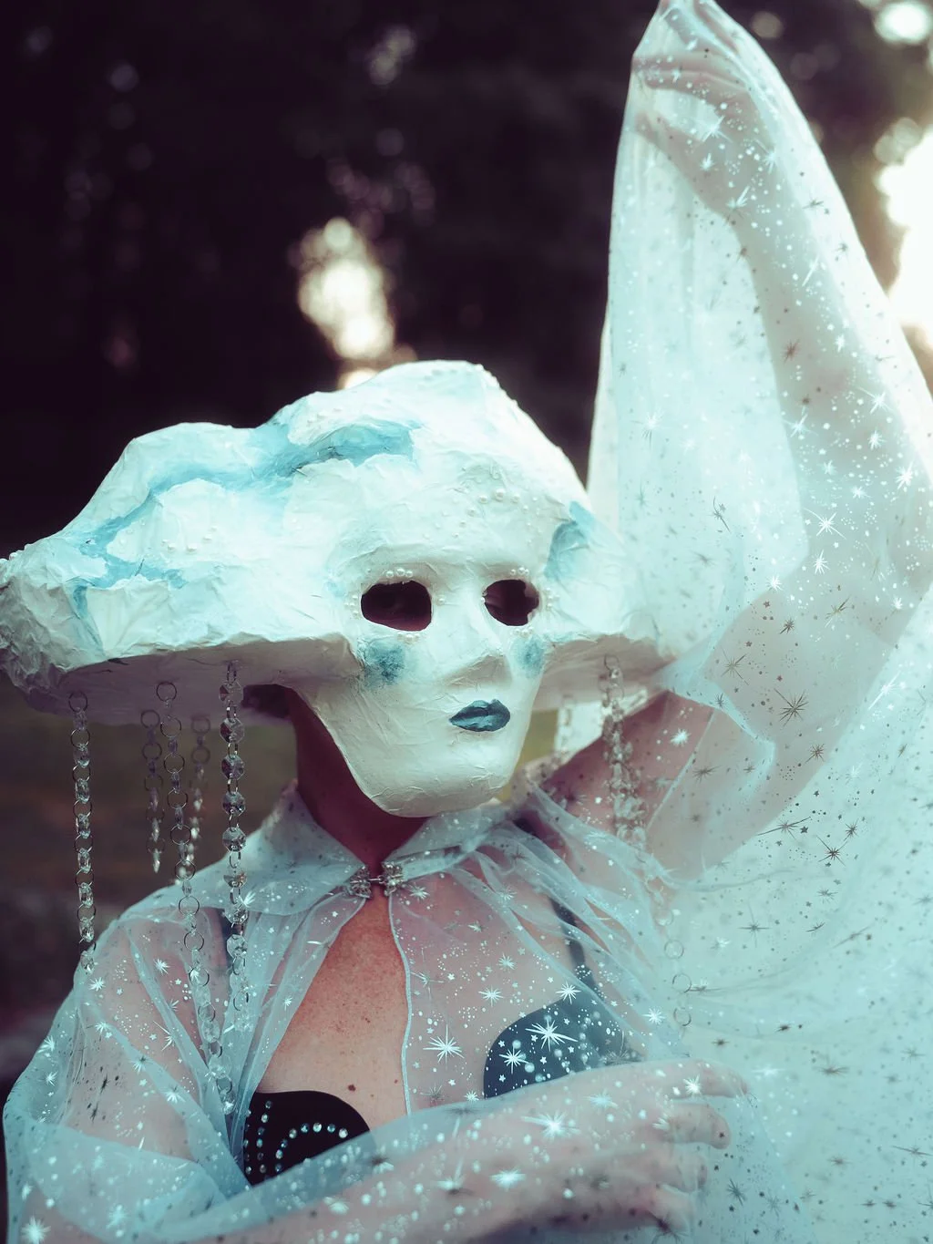 Person wearing a cartoonish skeleton mask and a large, white, cloud-like hat, with a sheer white cape with star patterns, standing outdoors.