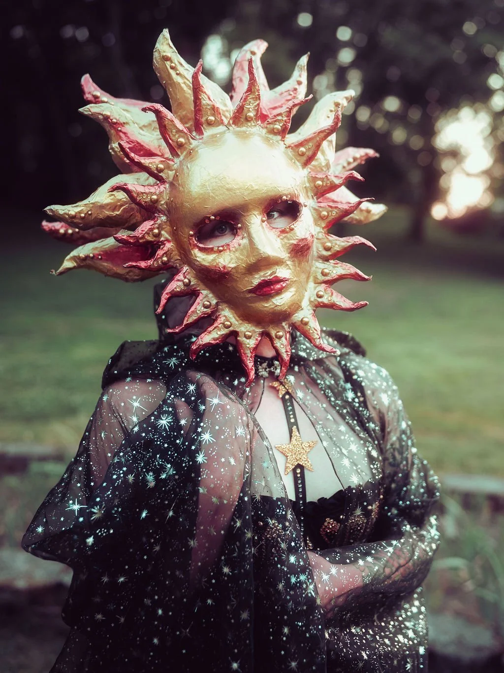 Person wearing a decorative sun-shaped mask with gold and pink accents, a star necklace, and a sheer black starry dress, outdoors during sunset or twilight.