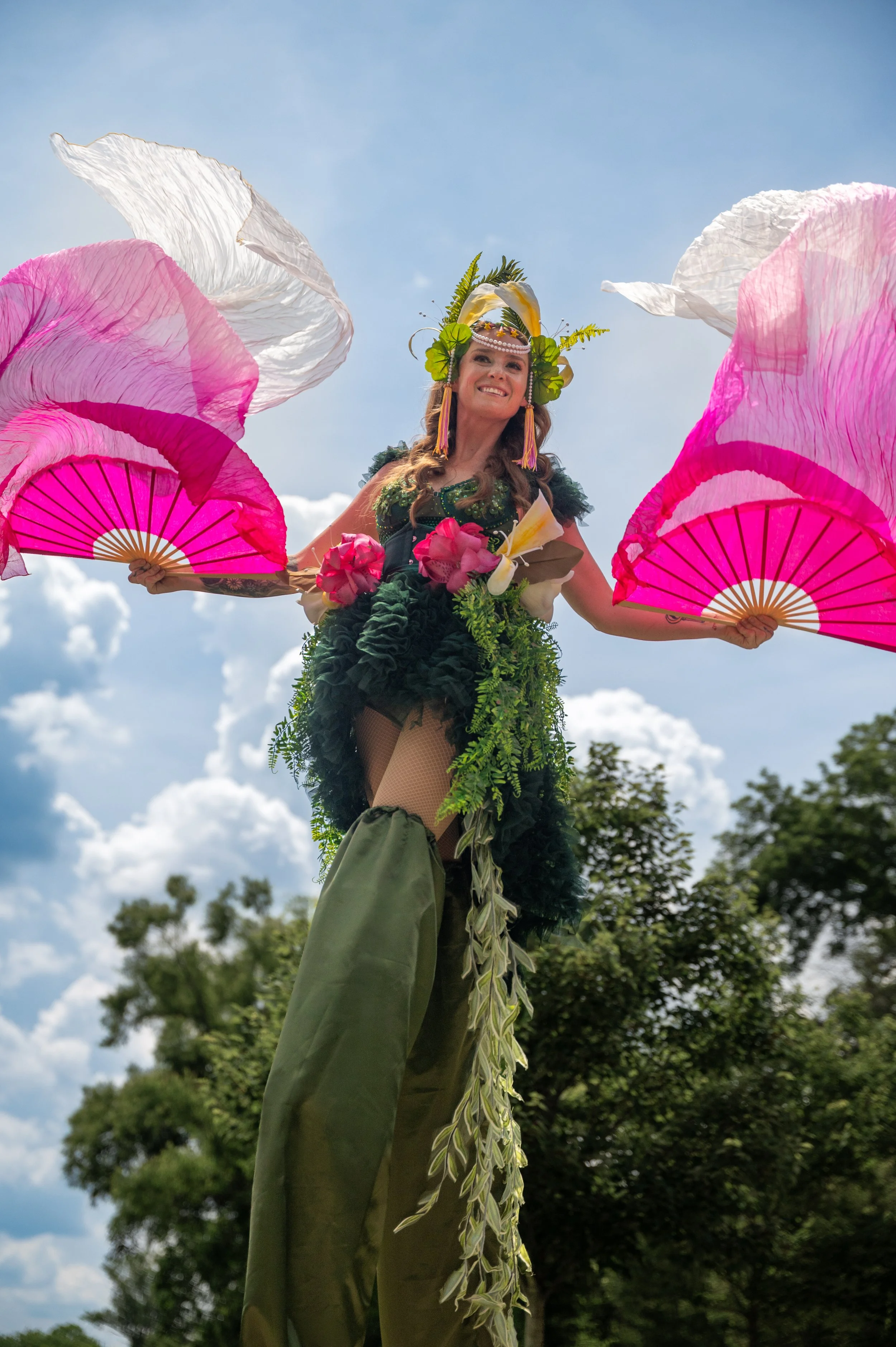 A woman dressed in a costume with greenery, holding pink and white fans, standing outdoors under a blue sky with clouds and trees in the background.