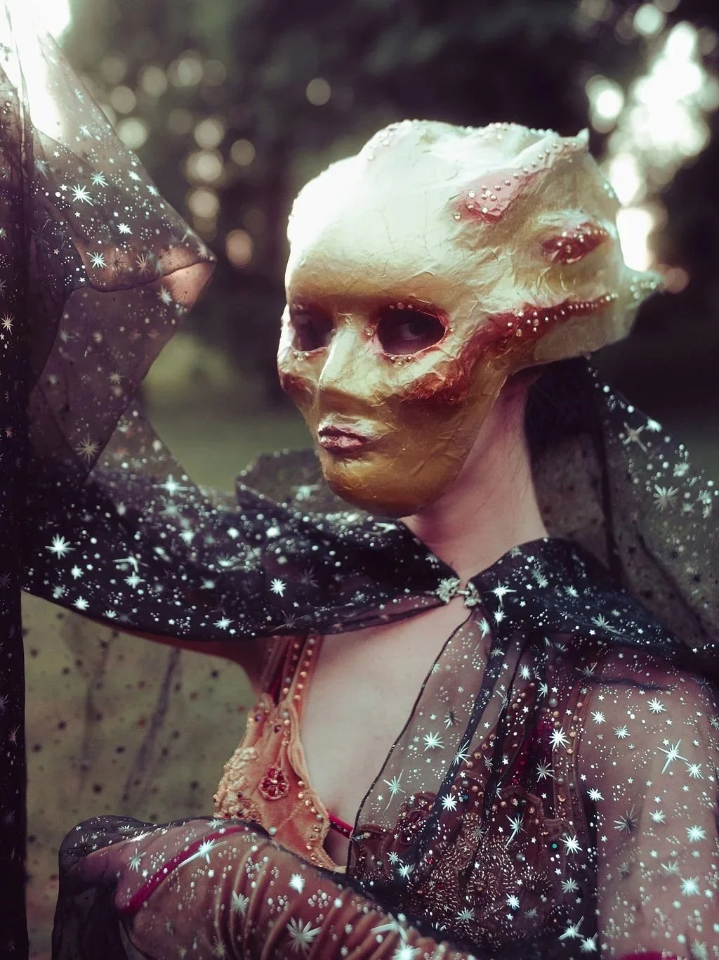 Person wearing a surreal, alien-like mask with a gold and flesh-toned appearance, decorated with small beads or pearls, and dressed in a black, starry, semi-transparent outfit with a cape.