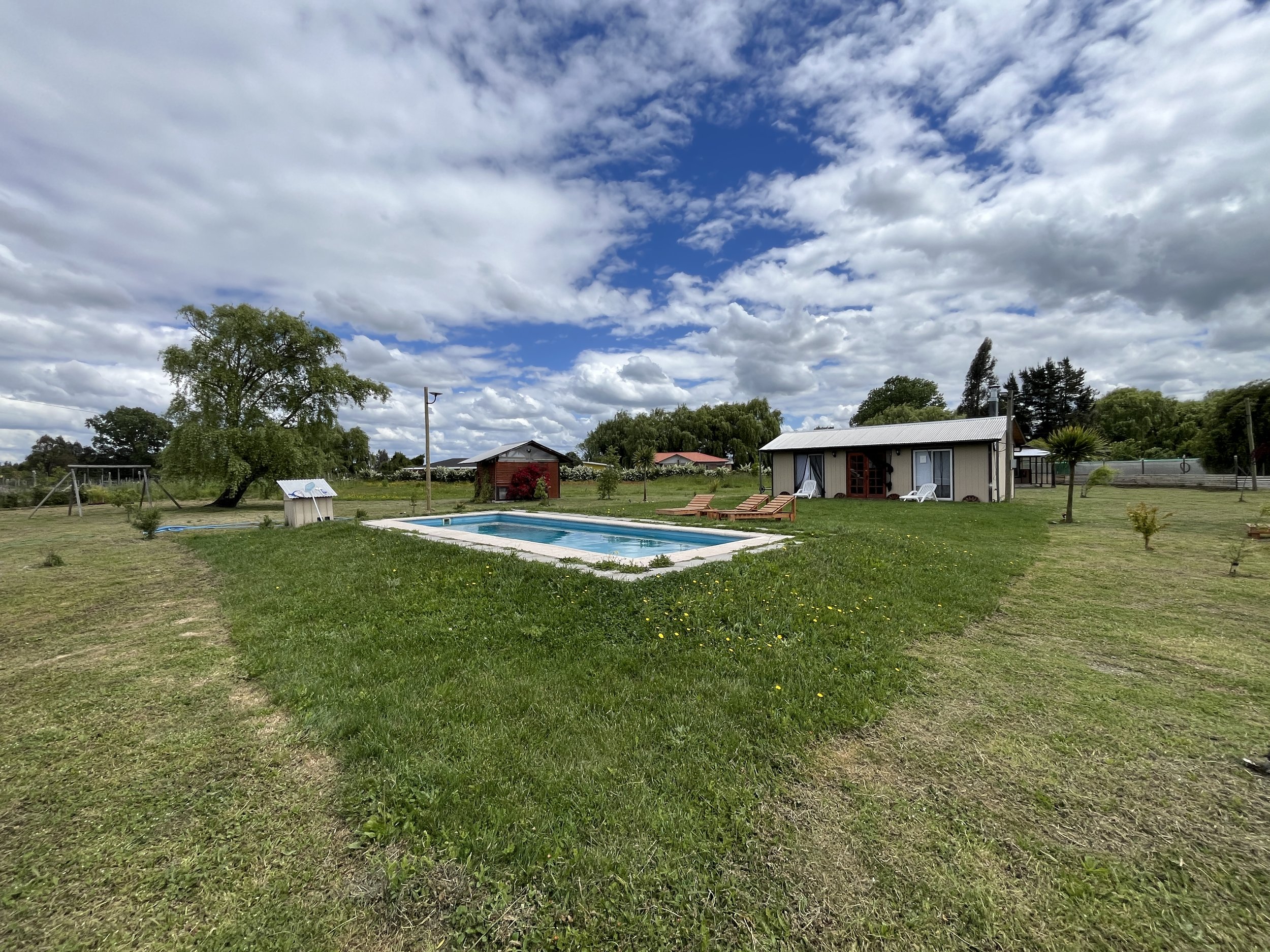 Patio con piscina, sillas y casas pequeñas en un día soleado con cielo parcialmente nublado.
