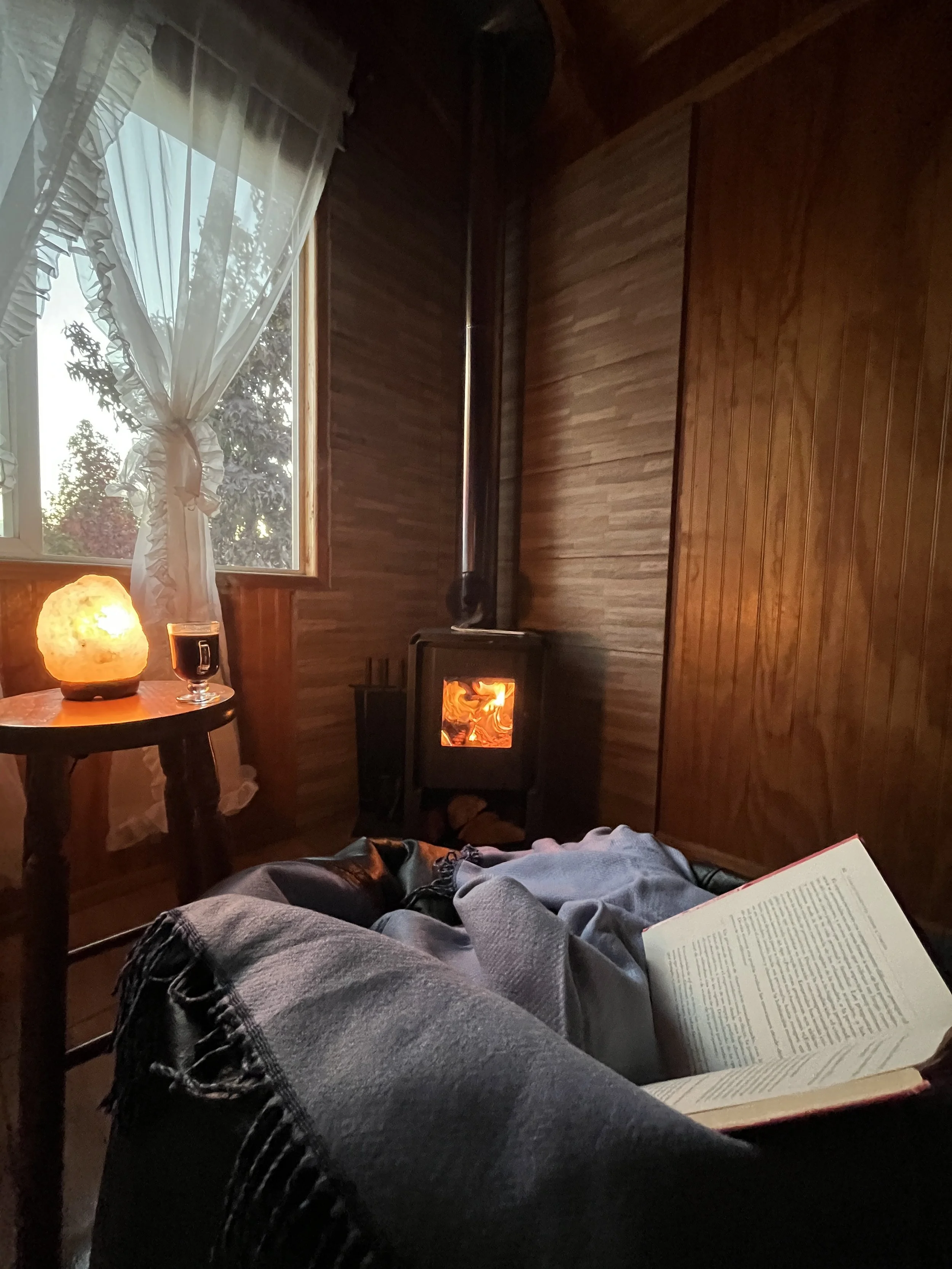 Una habitación con paredes de madera, una estufa de leña encendida, una mesa con una lámpara de sal, una ventana con cortinas y un libro abierto, observando desde una posición reclinada.