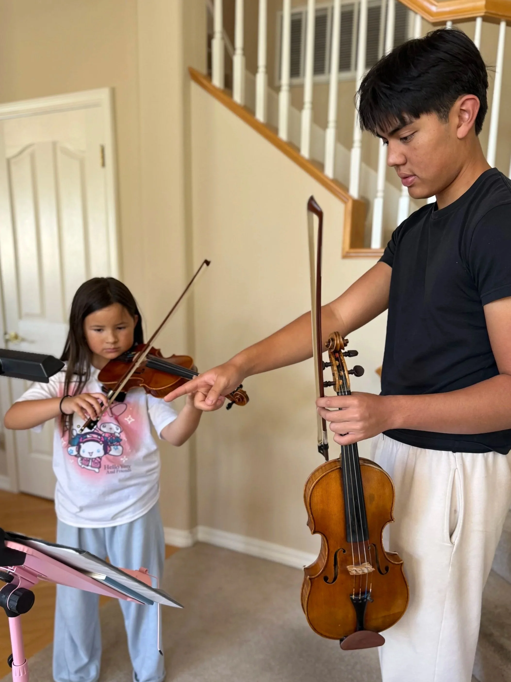 A young man and a young girl are playing violins together inside a house, with a staircase in the background.