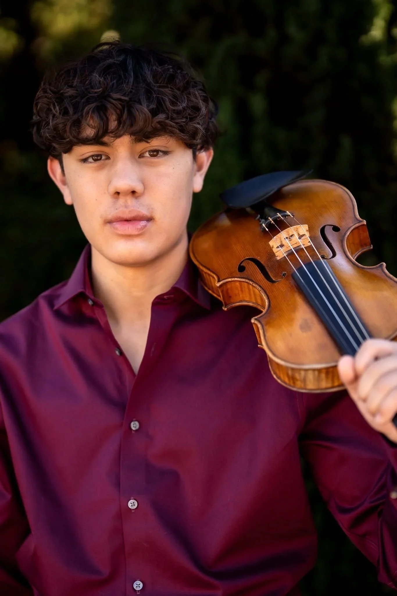 Young man with curly brown hair playing a violin outdoors, wearing a maroon button-up shirt, with a blurred green background.