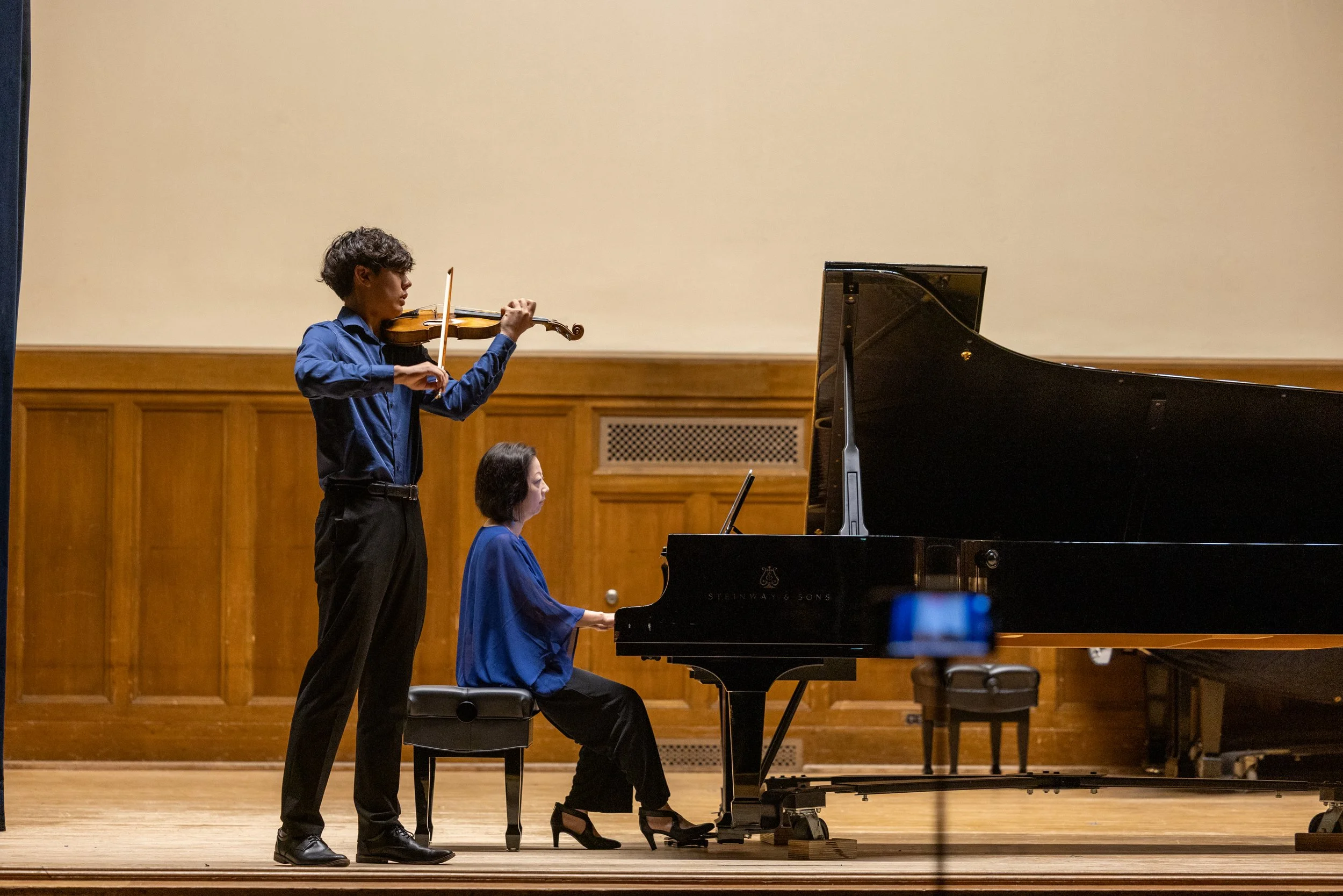 A young man playing the violin standing next to a woman playing the piano during a performance in a concert hall with wooden paneling.