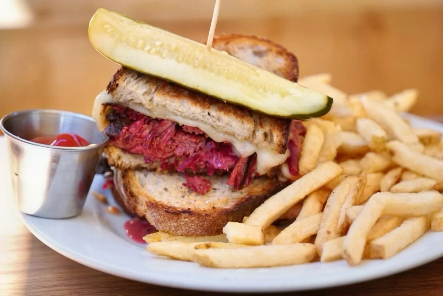 A close-up of a large sandwich with roast beef, melted cheese, toasted bread, topped with a pickle slice, served with French fries and a small cup of ketchup.