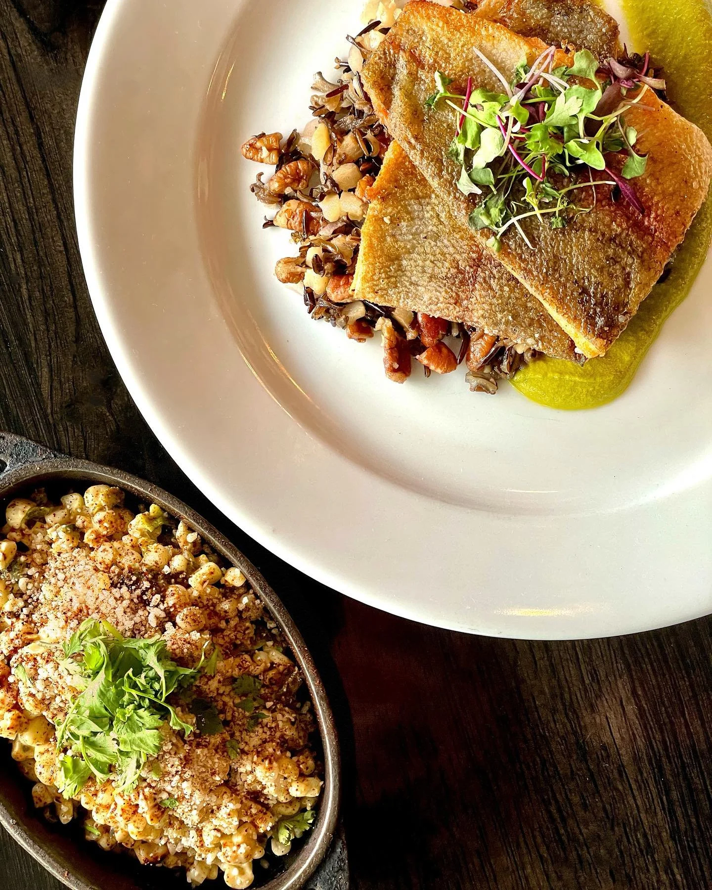 Close-up of a white plate with a cooked fish fillet topped with microgreens, served with a green sauce on the side, and black rice with vegetables and nuts in a skillet below.