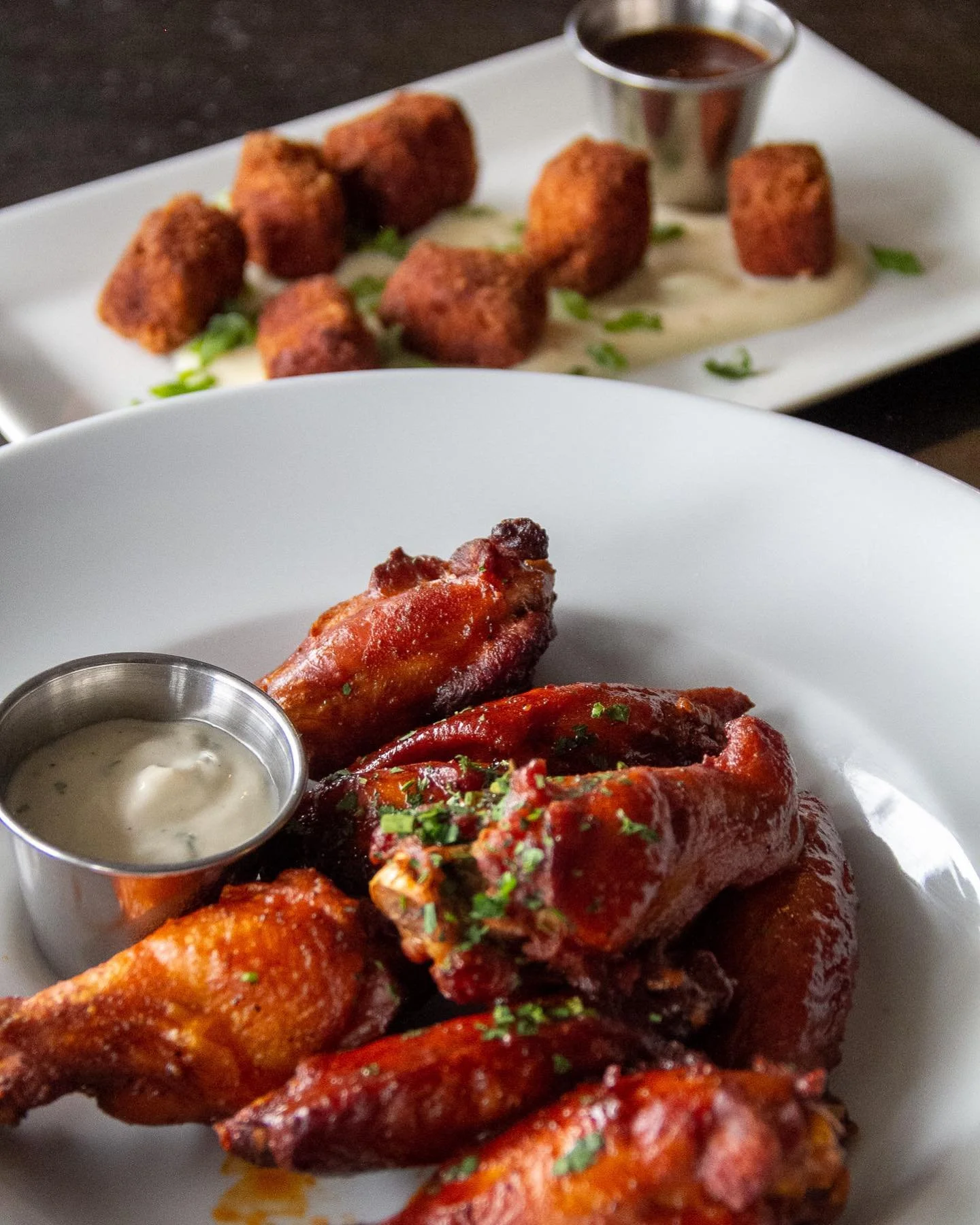 Close-up of spicy chicken wings garnished with chopped herbs, with a small cup of ranch dressing on the side, served in a white bowl.