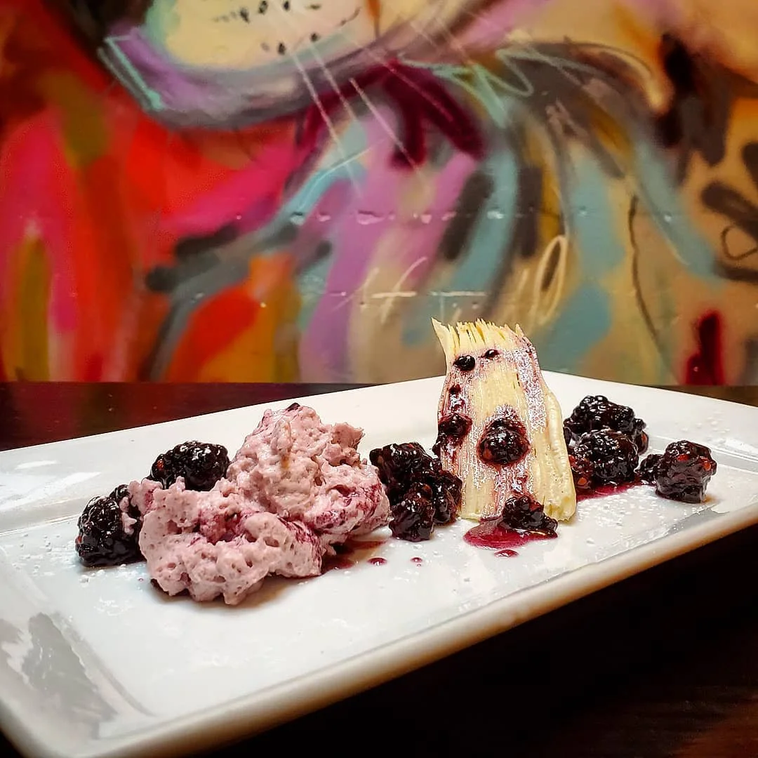 A plate with a scoop of pink berry ice cream, topped with blackberries and drizzled with berry syrup, along with a piece of vanilla ice cream with berry sauce on top, presented on a white rectangular dish, against a colorful, abstract background.