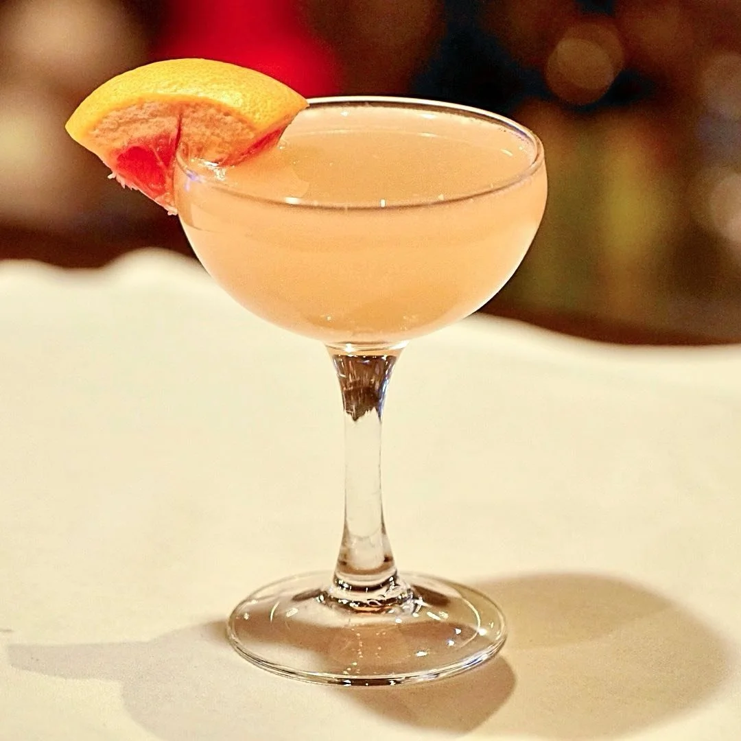 A cocktail glass with light pink beverage, garnished with a slice of lemon and grapefruit.
