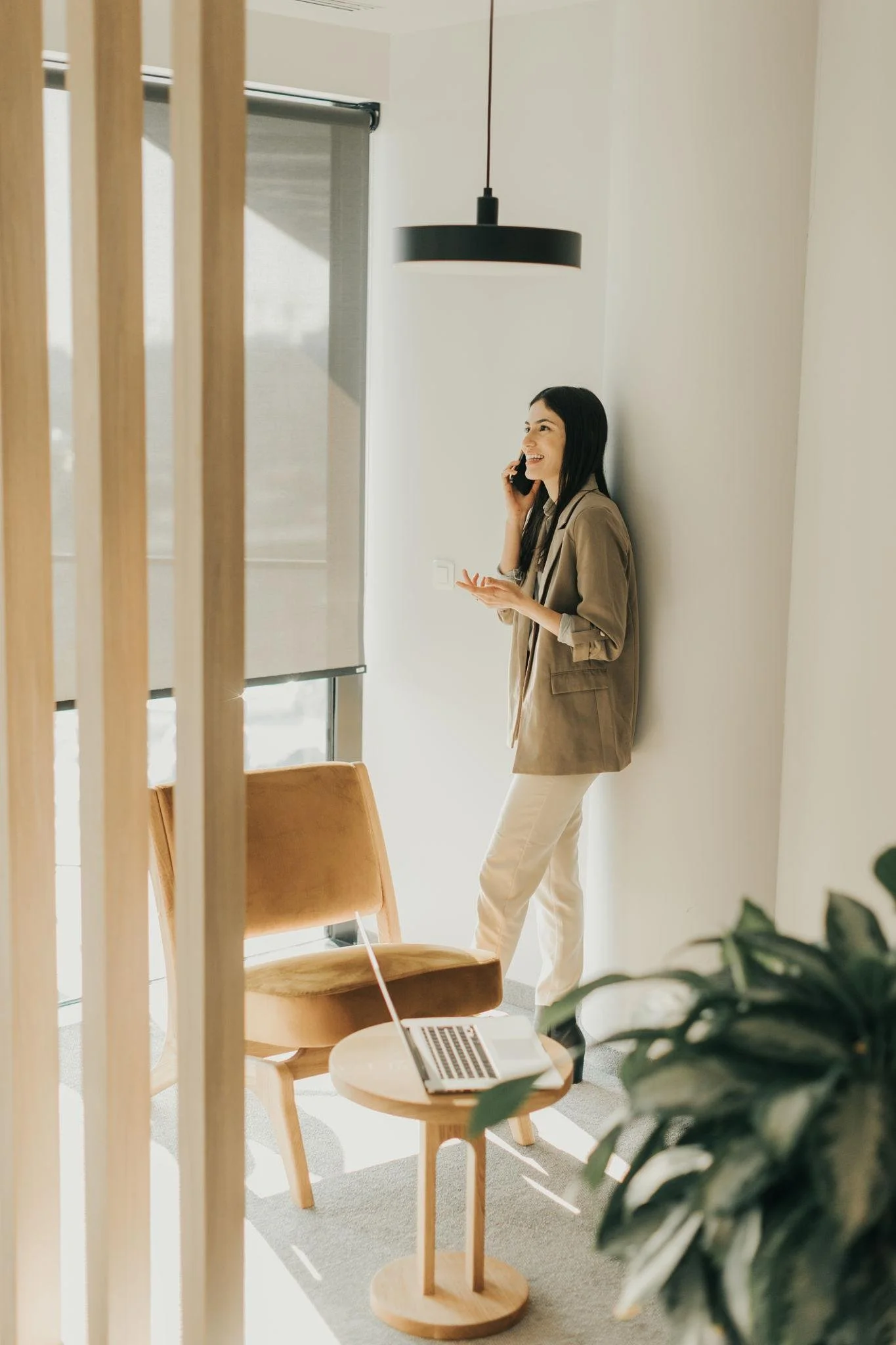 Woman standing in a bright modern room talking on the phone, with a laptop on a small round wooden table, and chairs nearby.