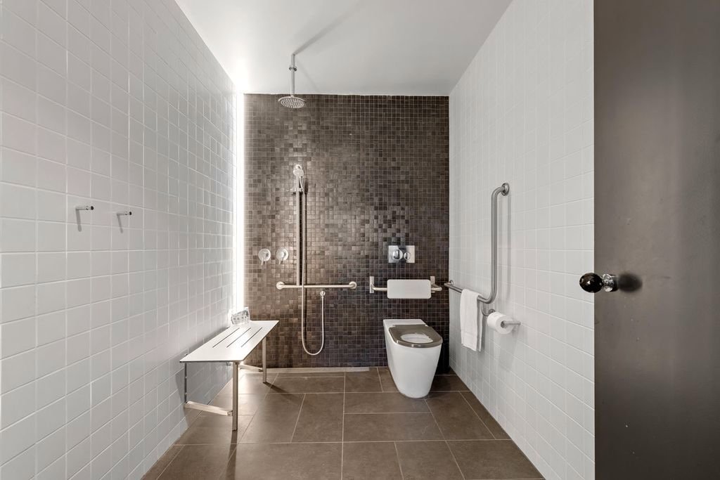 Accessible bathroom with a toilet, a shower area with a rainfall showerhead, grab bars on the wall, a towel rack, a small bench, and tiled walls in white and dark brown.