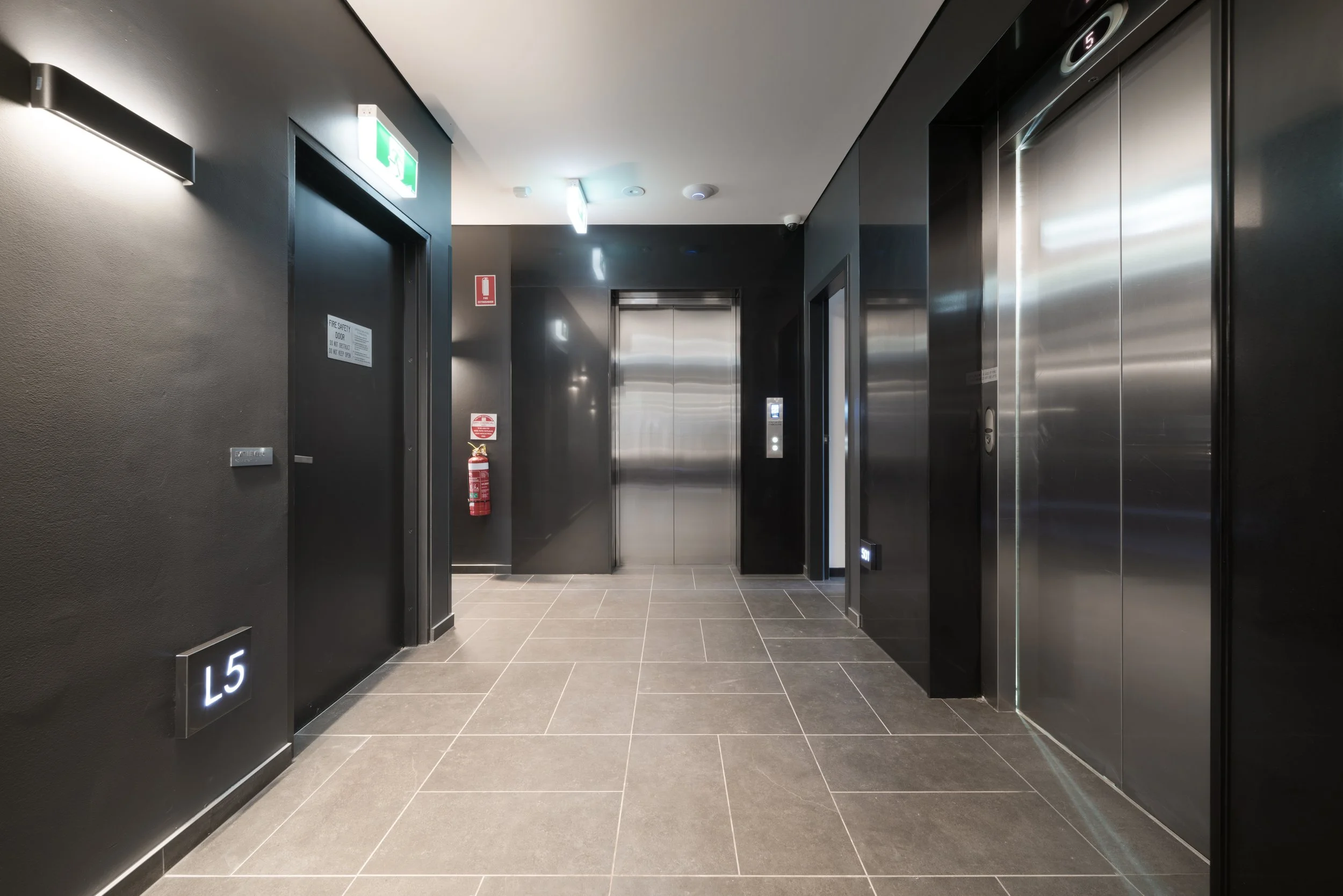 Elevator lobby with two elevators, black walls, light gray tiled floor, emergency fire extinguisher, exit signs, and a fire safety door.