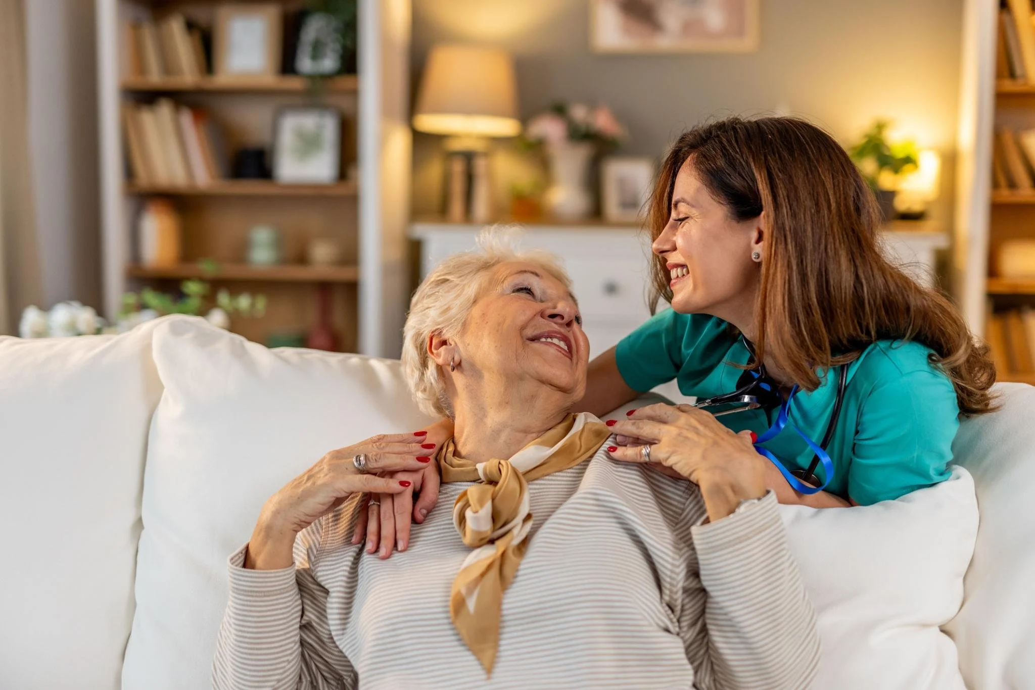 A healthcare worker smiling and talking to an elderly woman lying on a sofa in a warmly lit living room.