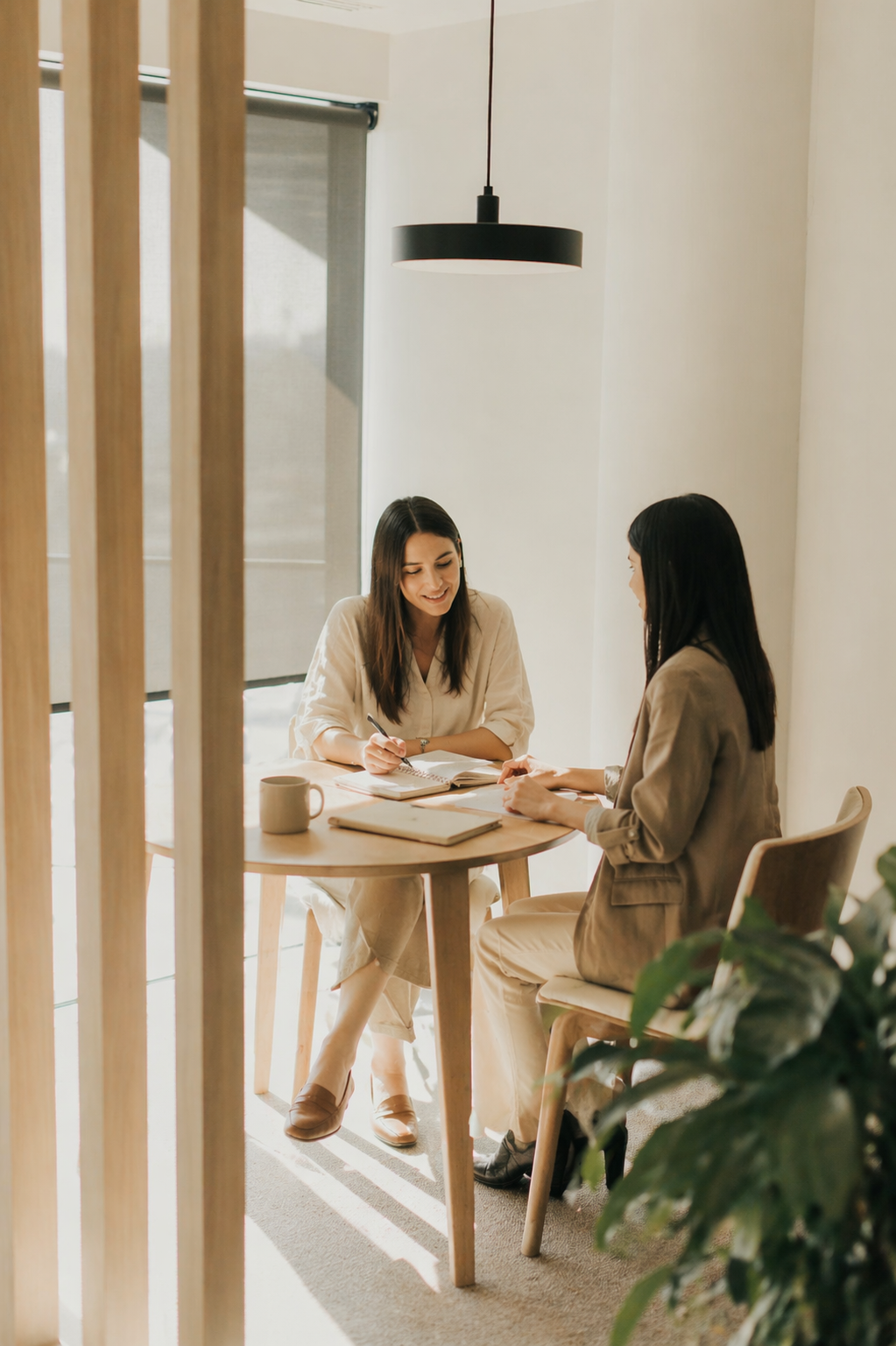 Two women sitting at a round wooden table in a bright, minimalist office, engaged in a conversation with notebooks, a coffee mug, and documents on the table.