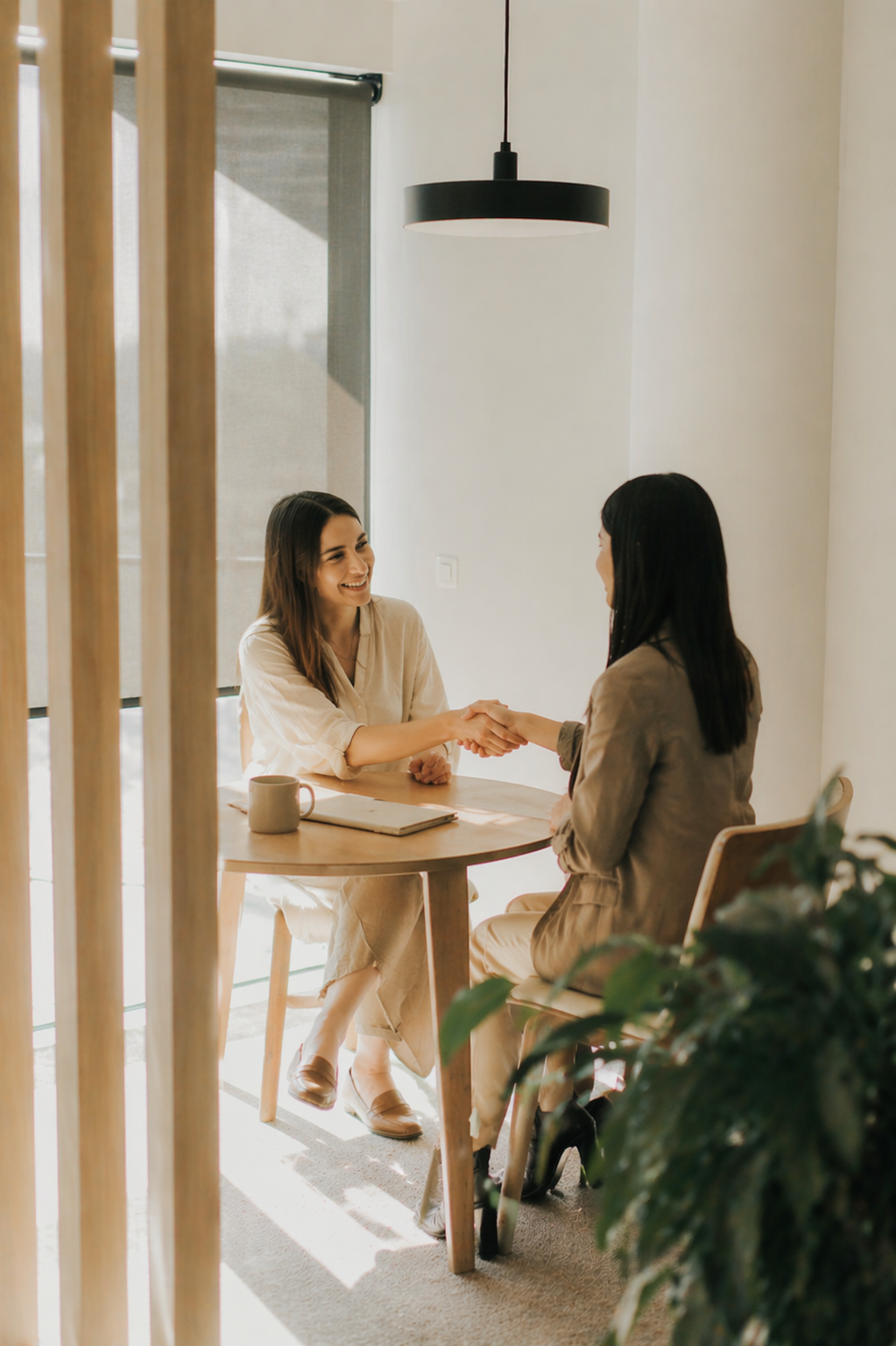 Two women shaking hands across a wooden table in a bright, modern room, smiling. One woman has brown hair and wears a beige outfit, the other has dark hair and wears a tan blazer. There is a coffee mug and a notebook on the table, with a large window and a black hanging light above.