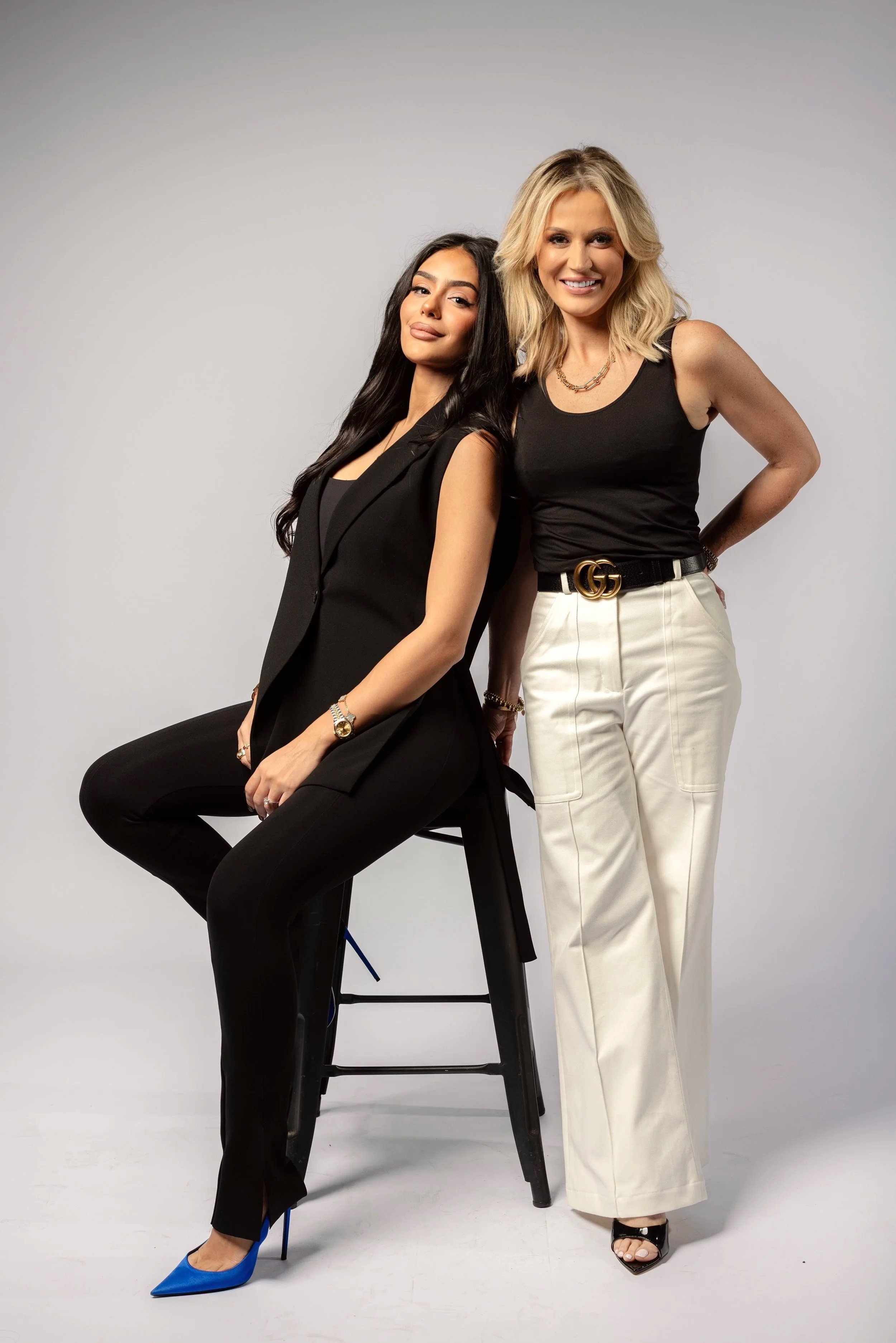 Two women posing in studio against a light gray background. One woman is sitting on a high stool, wearing a black sleeveless outfit and blue heels. The other woman is standing, dressed in white pants, a black sleeveless top, and black high heels with ankle straps. Both women are smiling and looking at the camera.