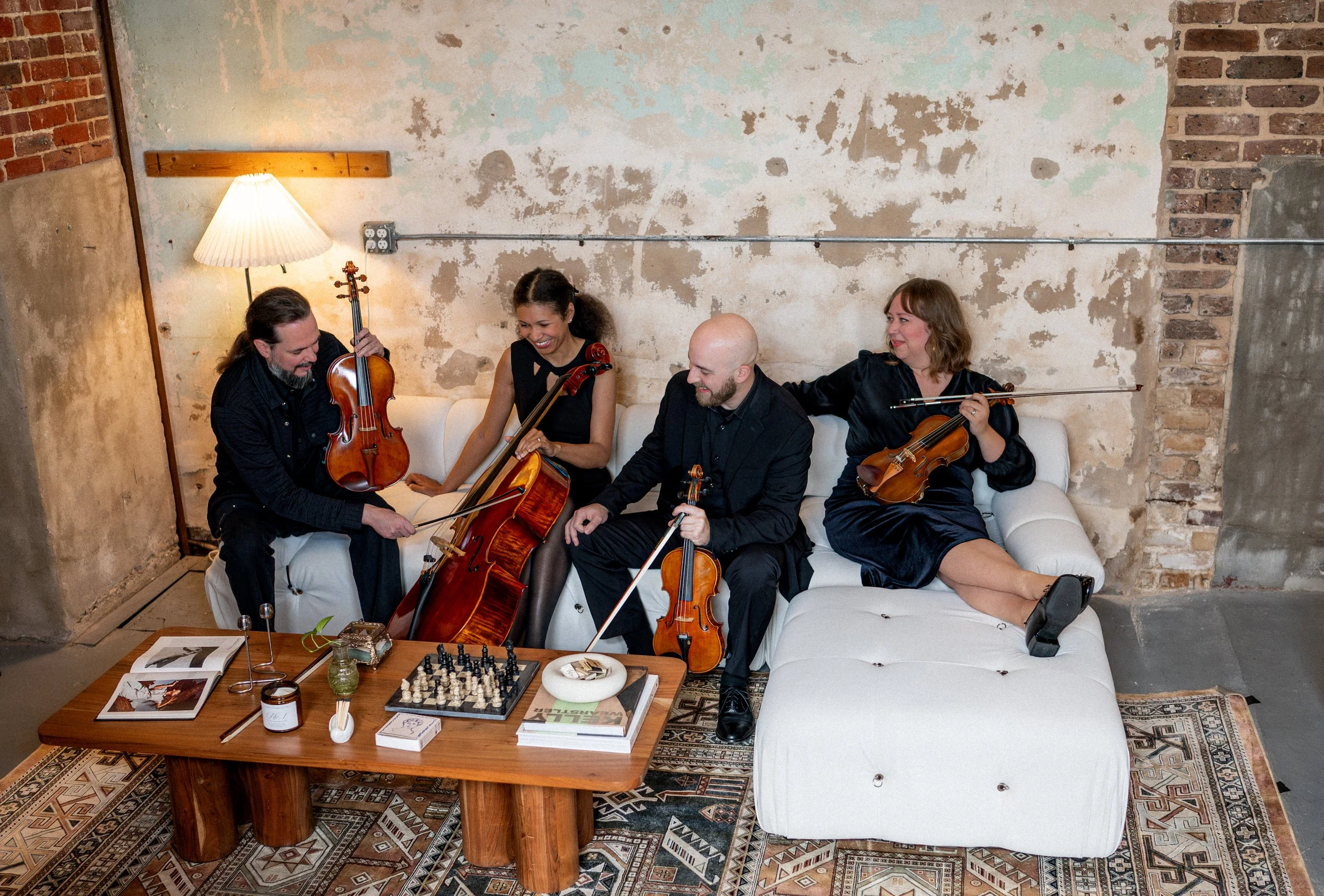 Group of four musicians playing string instruments in a cozy, rustic room with a worn brick and plaster wall.