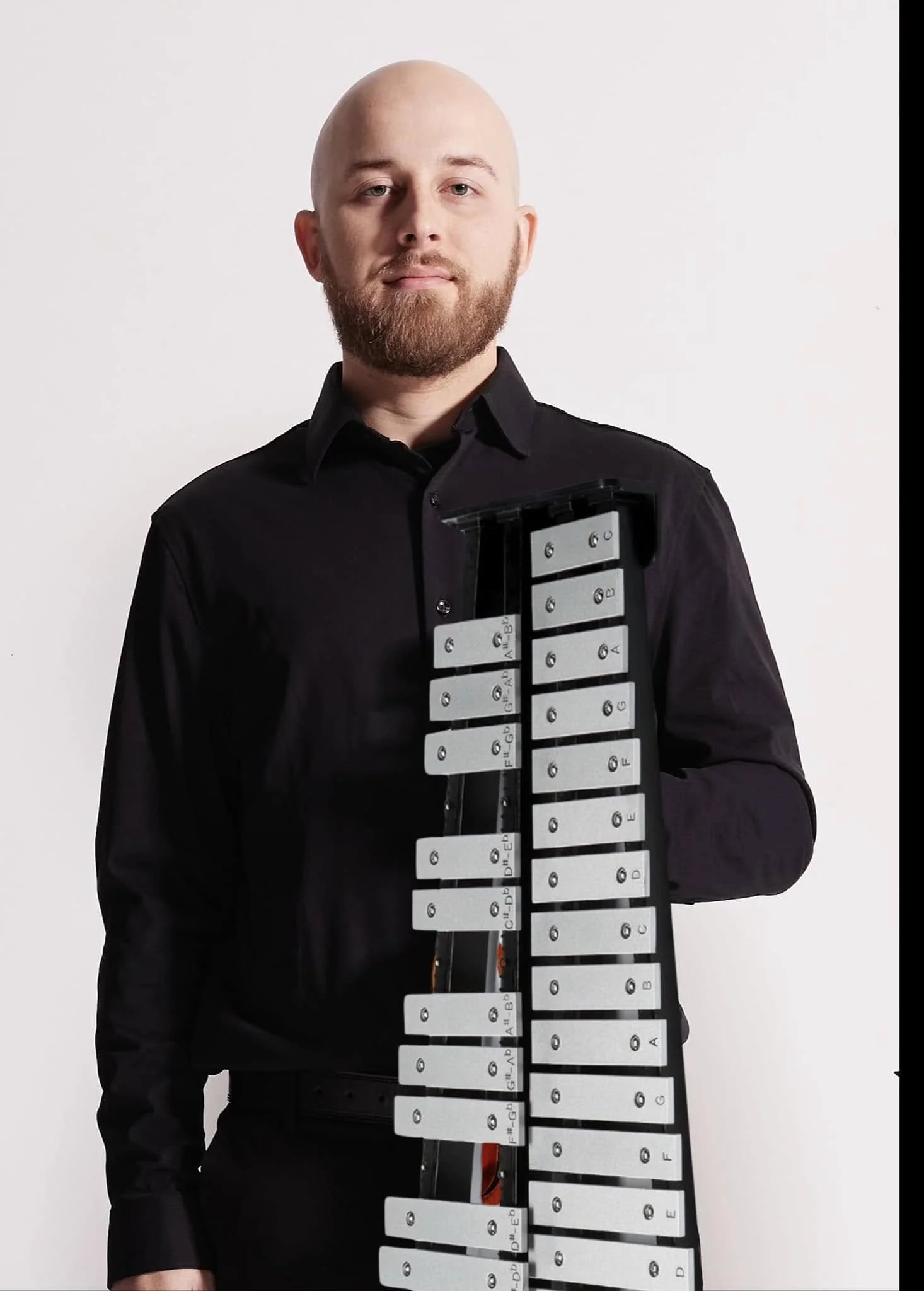 A man with a beard, wearing a black shirt, stands against a plain white background, holding a large xylophone with metallic bars labeled with musical notes.