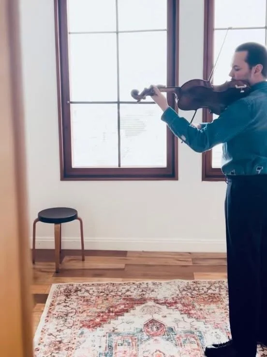 A man playing the violin in a room with large windows, a small stool, and a patterned rug on the wooden floor.
