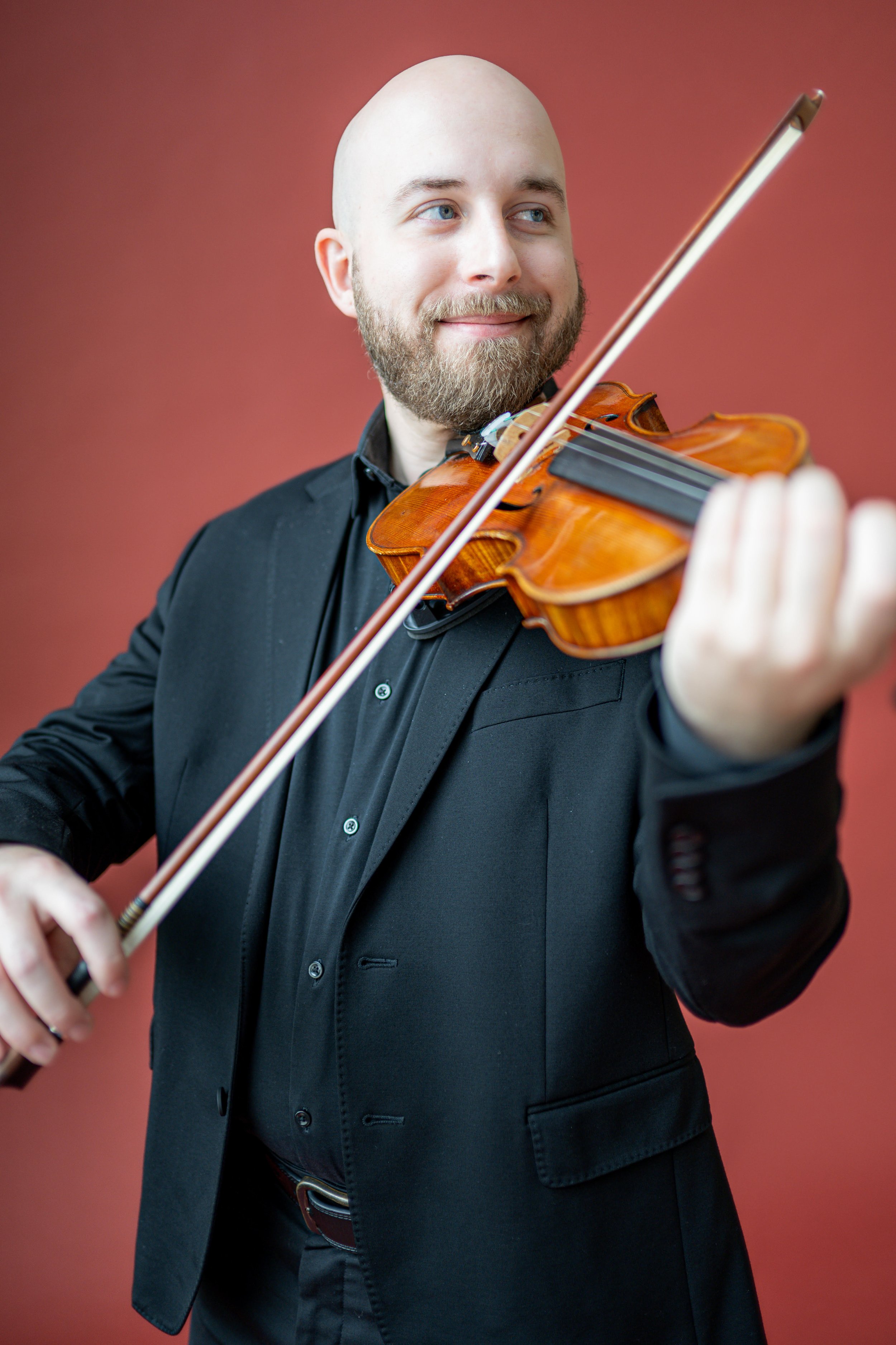 A man with a balding head and beard playing a violin and smiling, dressed in a black suit against a plain reddish background.