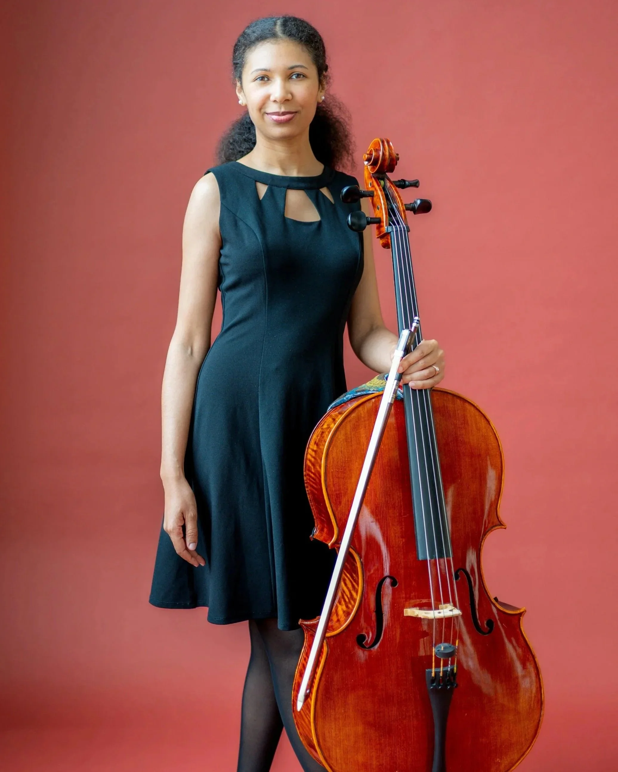 A woman with dark curly hair wearing a black dress and holding a cello standing against a pink background.