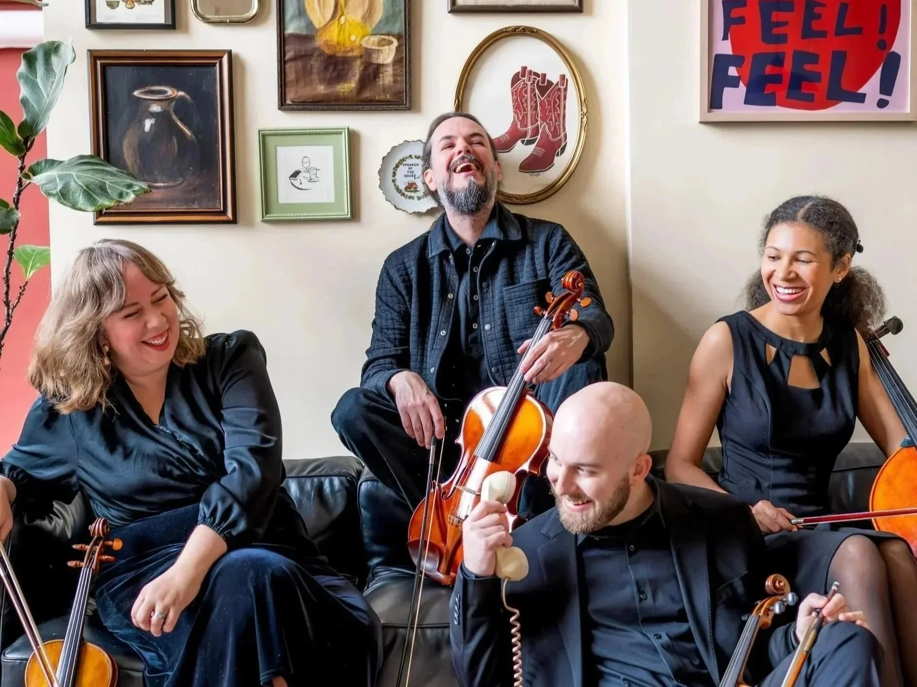 A group of five people enjoying music together in a cozy room with art on the wall. They are smiling and holding musical instruments like violins and a guitar, with one person playing the guitar and others playing violins.