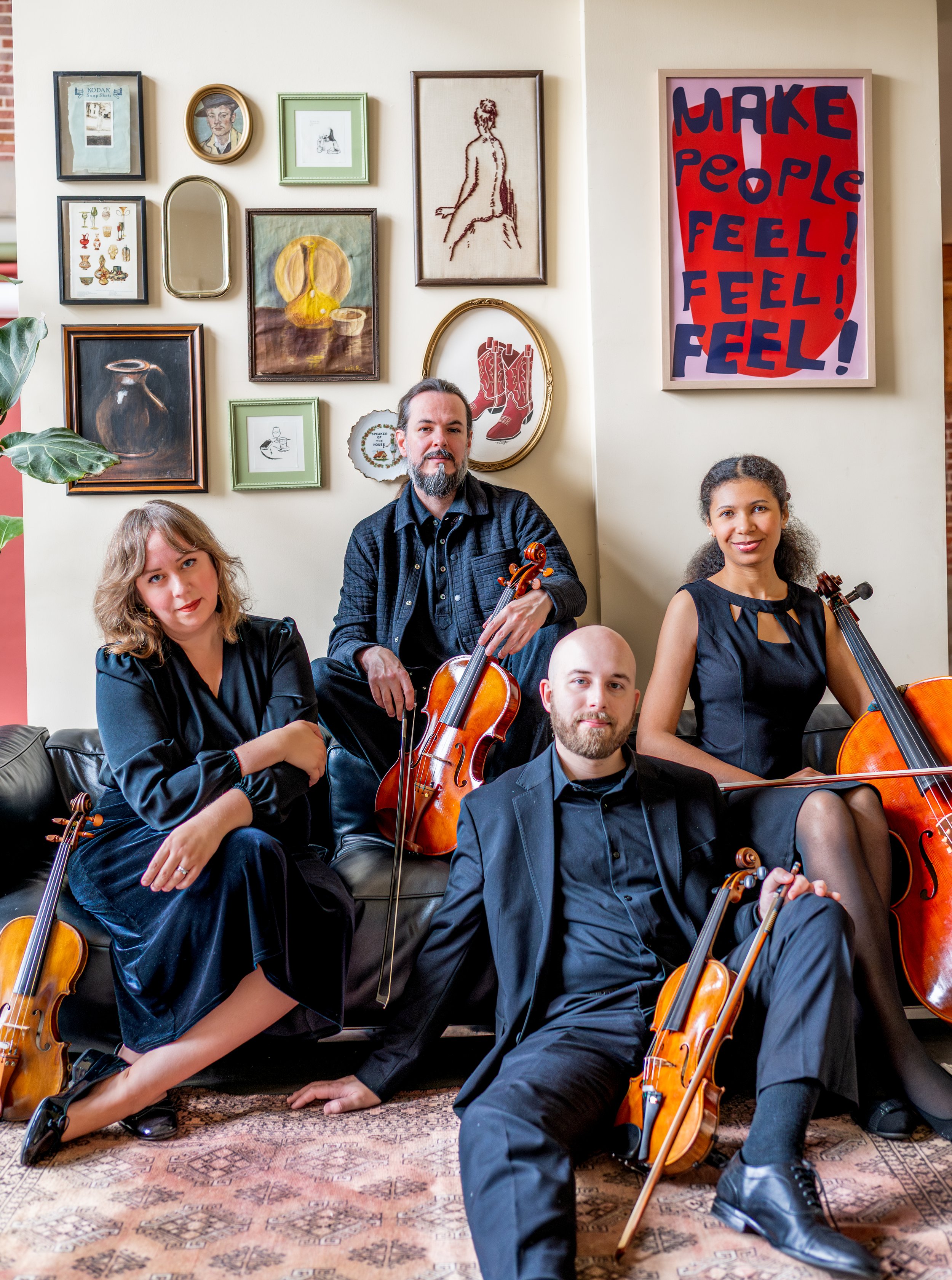 A group of five people posing with violins in a room with a wall decorated with framed artwork and a large poster that says 'MAKE PEOPLE FEEL! FEEL! FEEL!' The group is dressed in formal black attire, holding instruments and smiling at the camera.