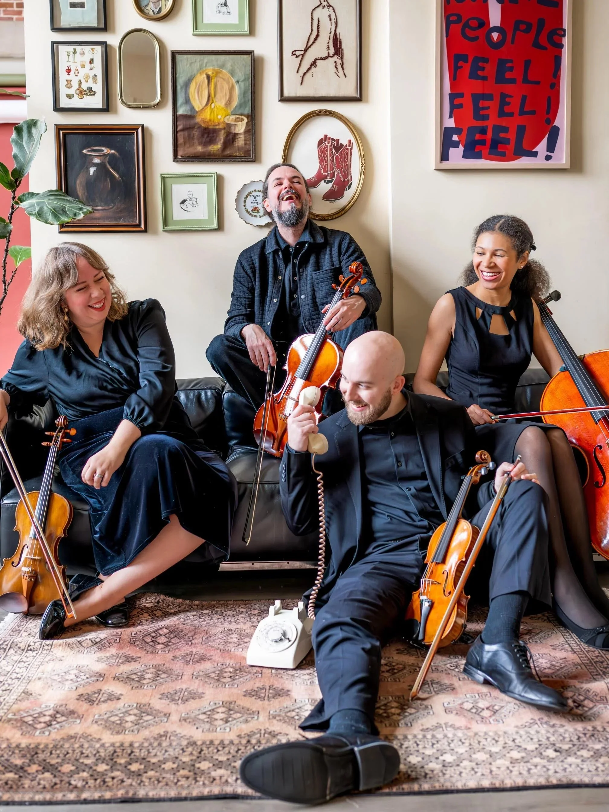 Five people sitting on a couch in a room with a gallery wall, smiling and playing musical instruments, including two violins and a cello. One person is talking on a vintage landline phone on the floor.