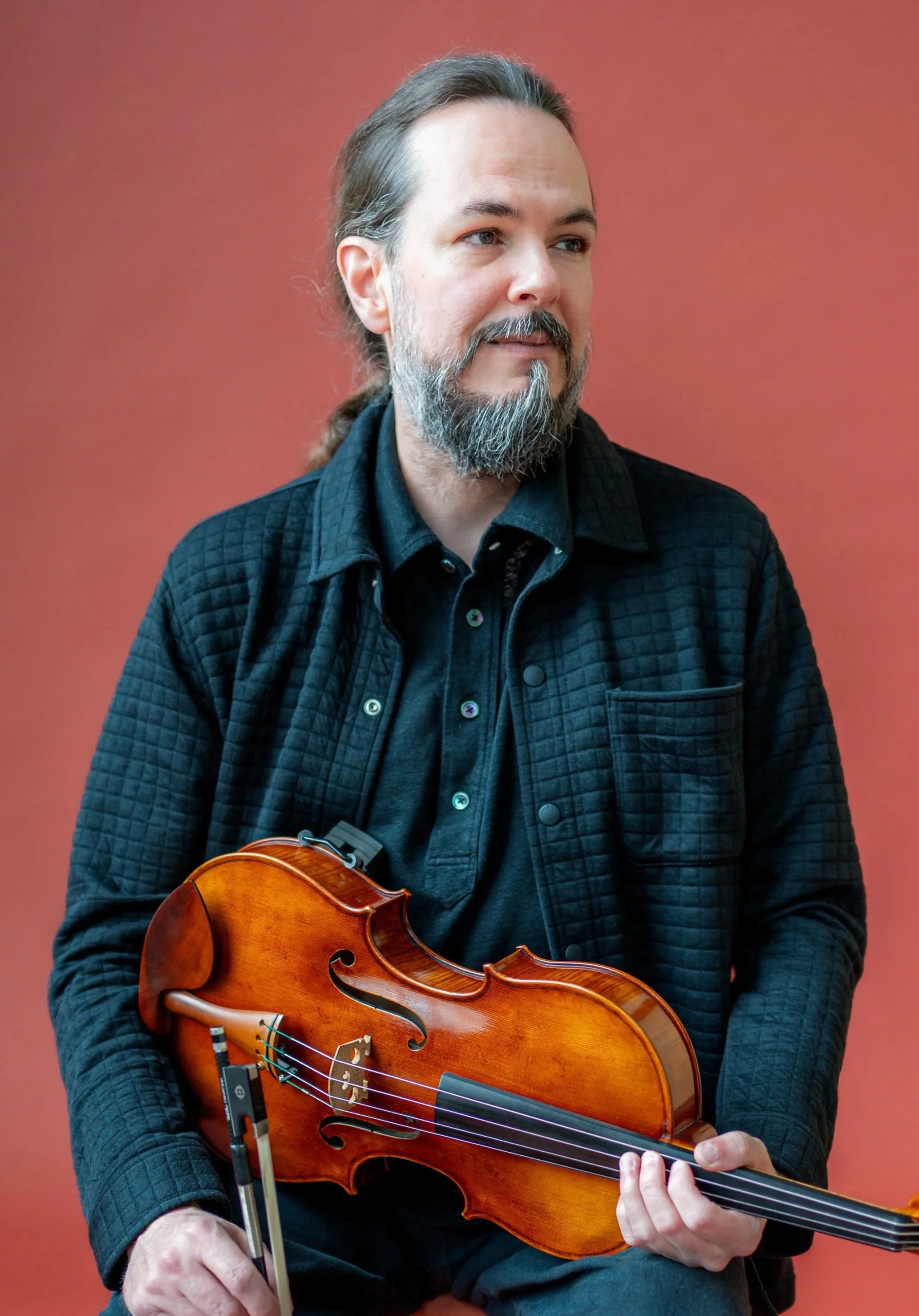A man with long gray hair and a gray beard playing a wooden violin against a solid red background.