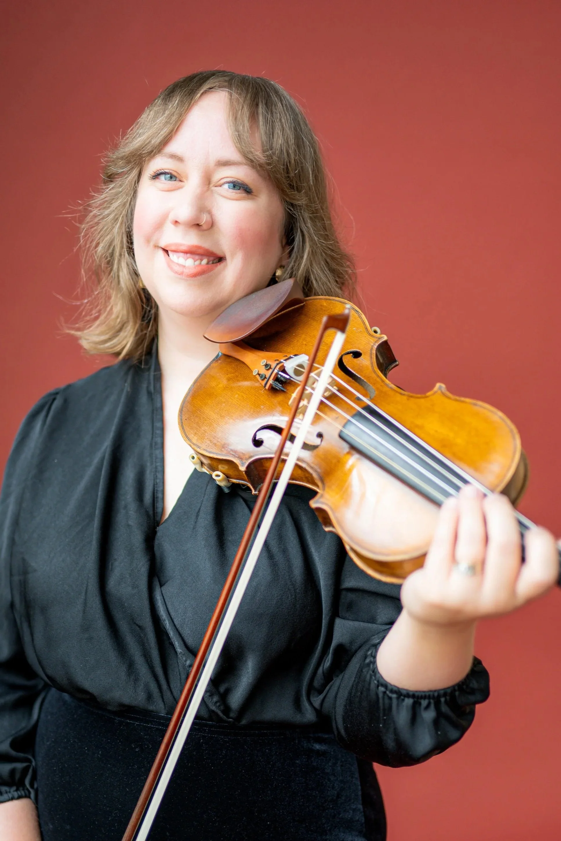 A young woman with light brown, wavy hair and blue eyes playing a violin, smiling at the camera, against a solid reddish-brown background.