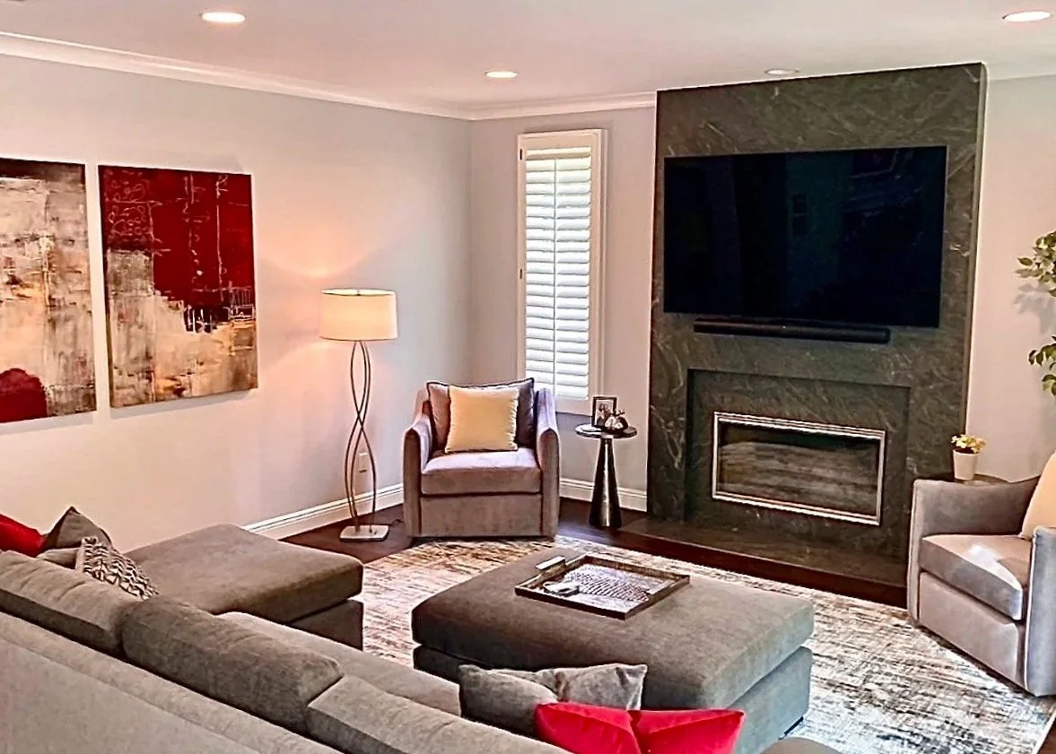Living room with gray sectional sofa, two armchairs, a fireplace with a large TV mounted above, a floor lamp, wall art, window with shutters, and various small decorative items.