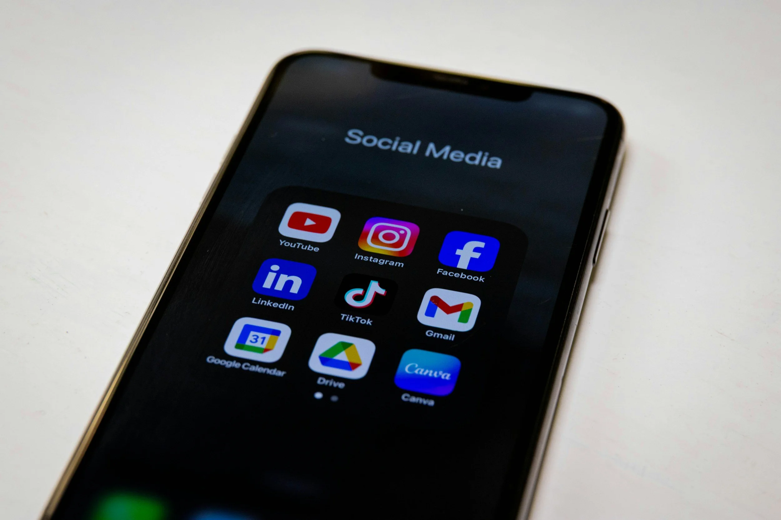 Close-up of a smartphone screen displaying social media app icons such as YouTube, Instagram, Facebook, LinkedIn, TikTok, Gmail, Google Calendar, Drive, and Canva on a white surface.