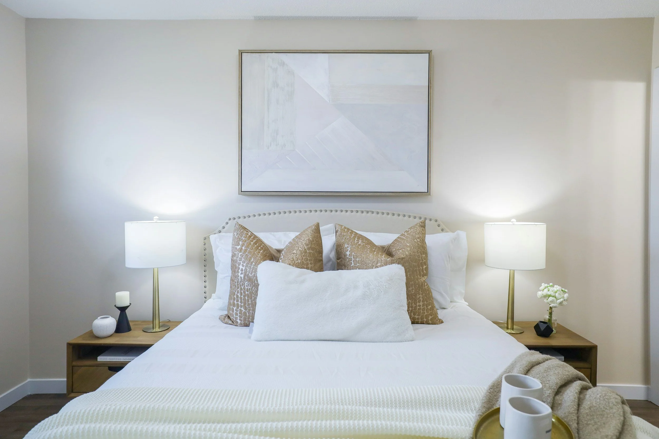 A neatly made bed with beige and white pillows, flanked by two matching bedside tables with lamps, a large abstract painting hanging above the headboard, and decorative vases on the tables, in a well-lit bedroom with light-colored walls.