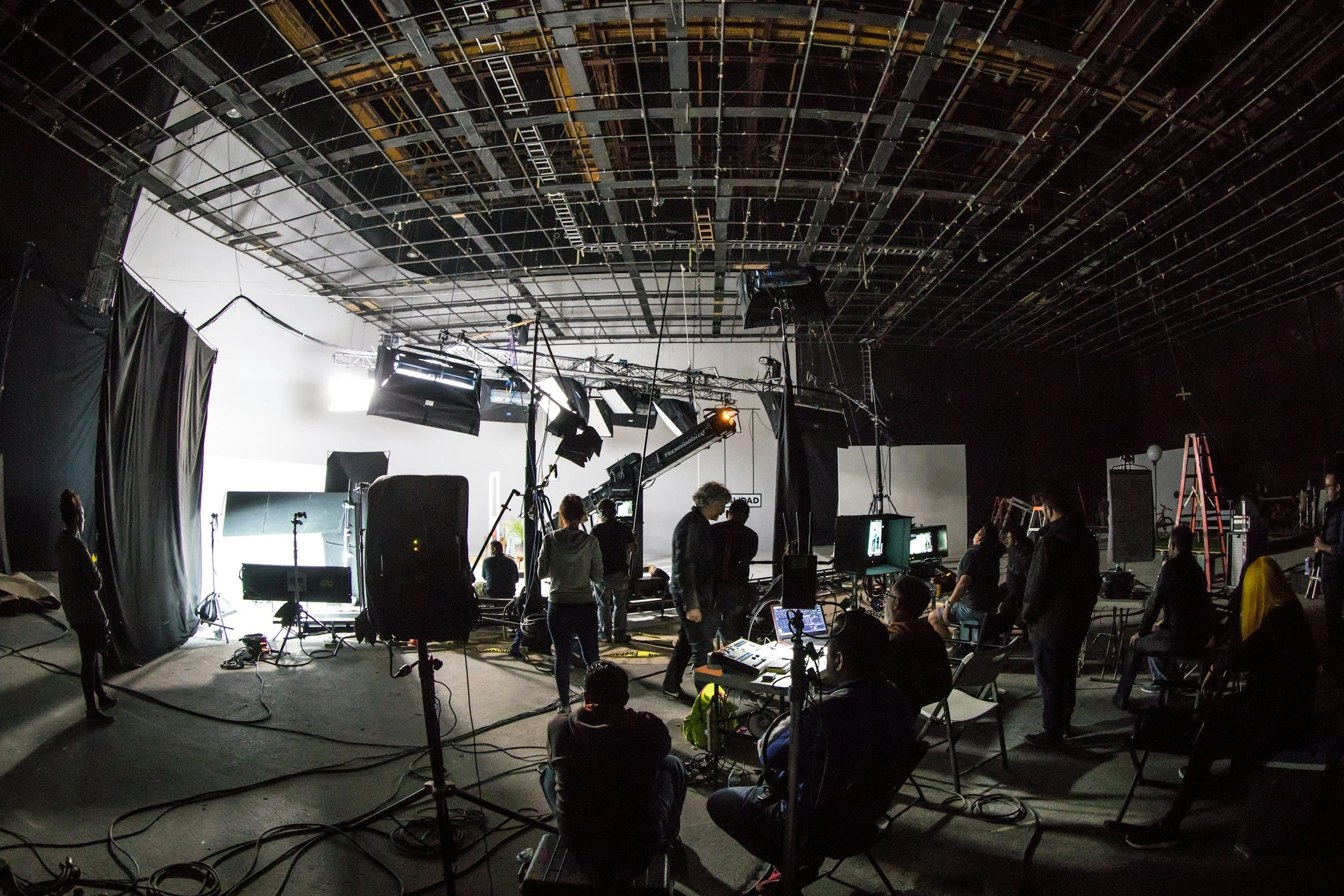 View of a film or television studio set with crew members preparing equipment and lights, with a white backdrop and a grid of rigging overhead.