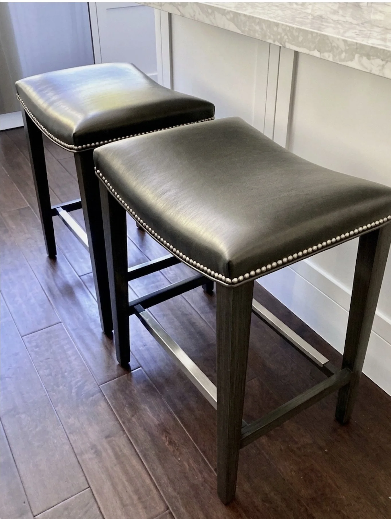 Black upholstered barstool with nailhead trim, on dark wooden legs, placed in front of a white bar counter with a marble top, on a dark wood floor.