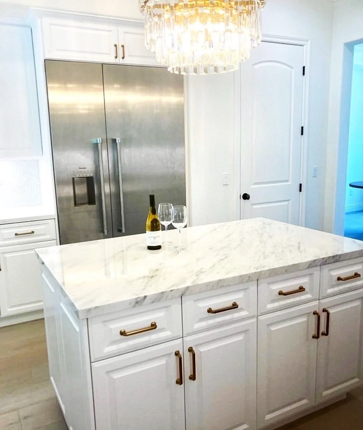 A kitchen with a marble island, a large stainless steel refrigerator, white cabinets, and a chandelier hanging above the island. There is a bottle of wine and two wine glasses on the marble surface.