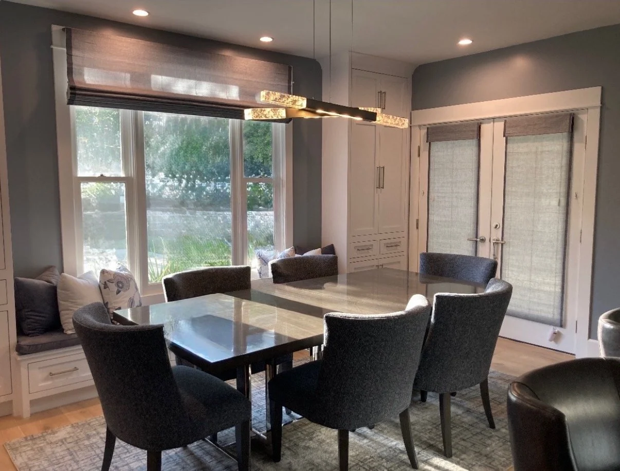 A dining room with a rectangular table surrounded by six dark gray upholstered chairs, a window seat with pillows beneath large front windows, a built-in white cabinet, and double doors with shades. Modern pendant lights hang above the table, and recessed ceiling lights are installed. The room has gray walls and hardwood flooring.