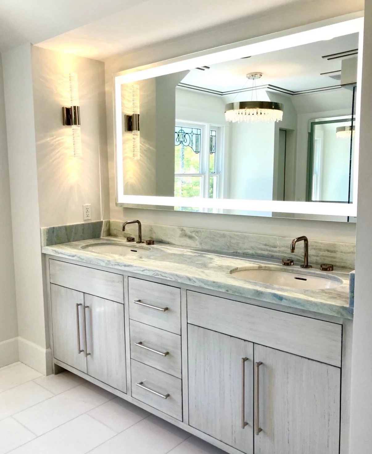 Modern bathroom vanity with marble countertop, double sinks, large backlit mirror, and stylish wall sconces, reflected in a large mirror.