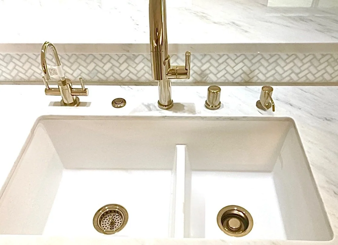 A white kitchen sink with gold fixtures, including a gooseneck faucet, a smaller faucet, and a soap dispenser, set in a marble countertop with a decorative backsplash.