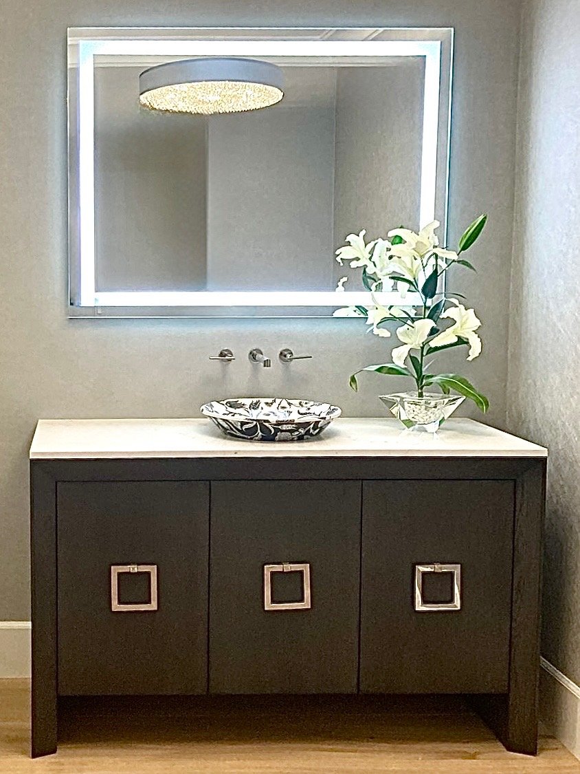 Modern bathroom vanity with a decorative bowl and a vase with white lilies, illuminated mirror, and dark cabinet with square handles.