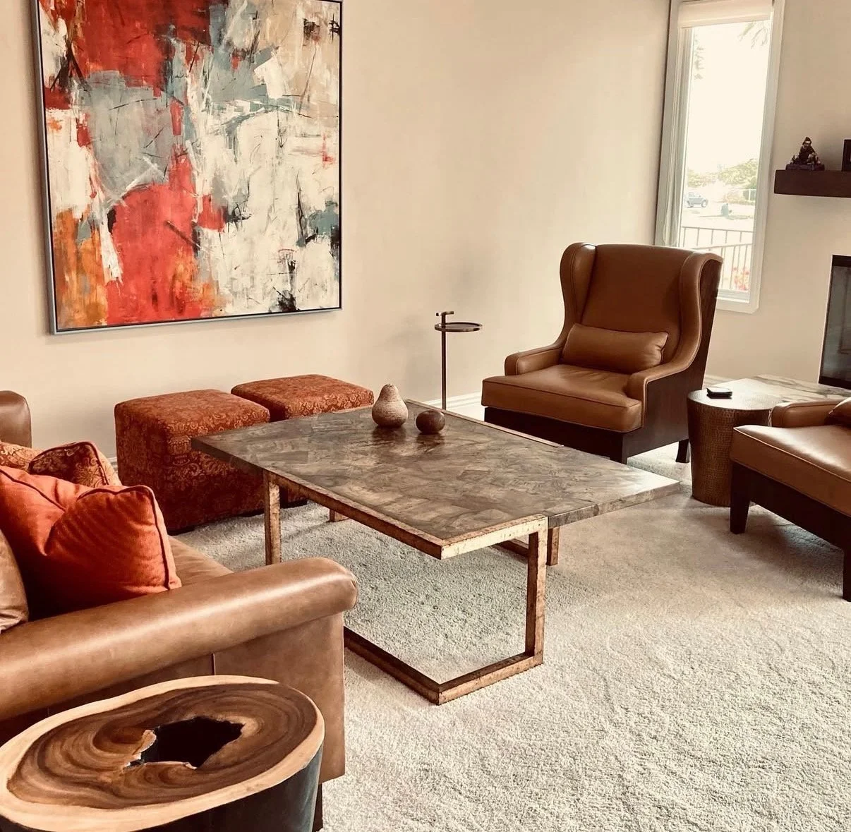 Living room with abstract art on the wall, brown leather armchairs, a marble coffee table with decorative vases, two red patterned ottomans, a window with view outside, and a small side table with a TV remote.