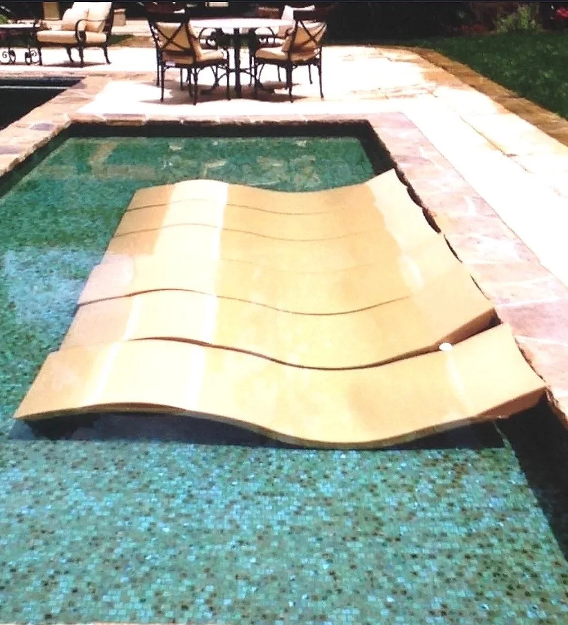 A backyard swimming pool with a yellow pool float shaped like a smiley face in the water. There is a brick patio surrounding the pool with outdoor chairs and a table in the background.