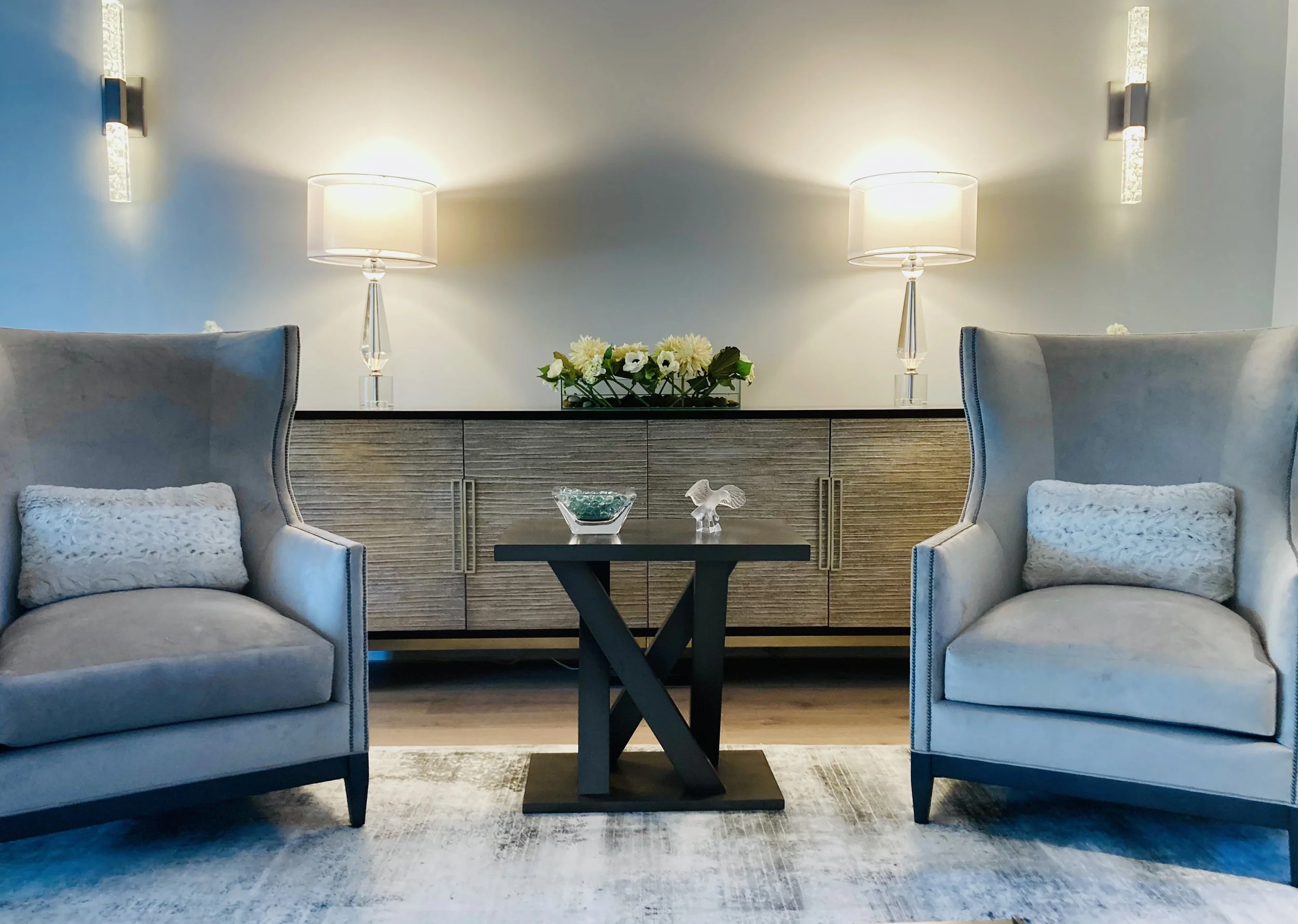Living room with two armchairs, a black coffee table, and a sideboard with two lamps and floral decorations. The room has a light, neutral color scheme and modern decor.