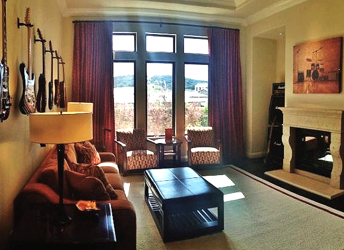 Living room with four guitars on the wall, a sofa with pillows, a floor lamp, a coffee table, two armchairs by the window, a fireplace, a painting above the fireplace, and large windows with curtains.