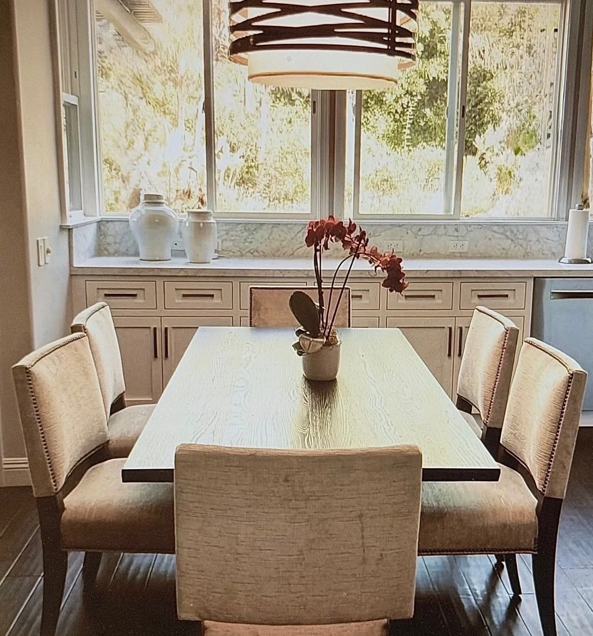 A dining room with a rectangular wooden table, six beige upholstered chairs, a potted orchid in the center, large windows with trees outside, a pendant light hanging above, and white vases on the sideboard.