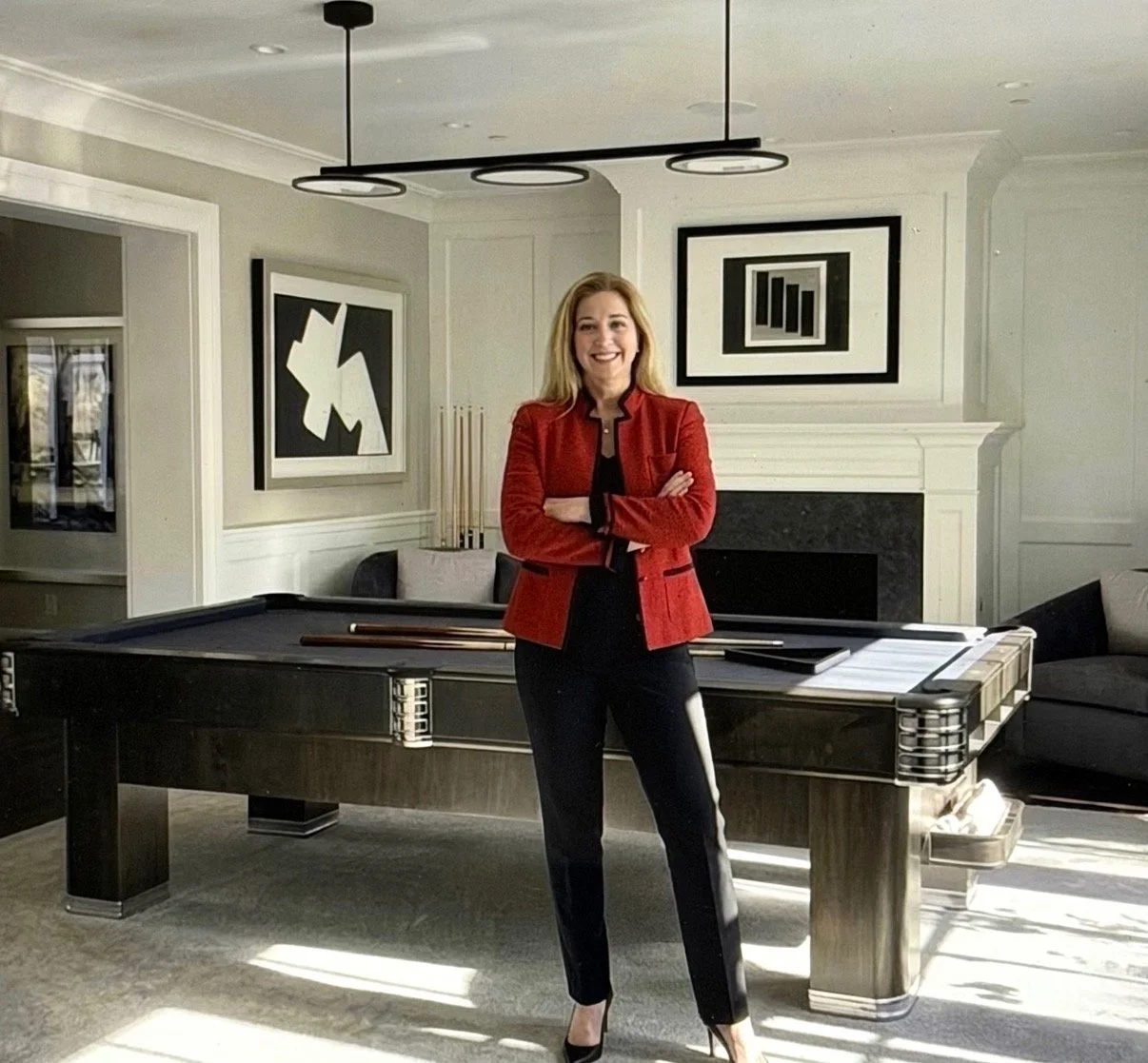 Woman with blonde hair wearing a red blazer and black pants standing with arms crossed in a modern living room with a pool table, abstract black and white artwork, and a fireplace.